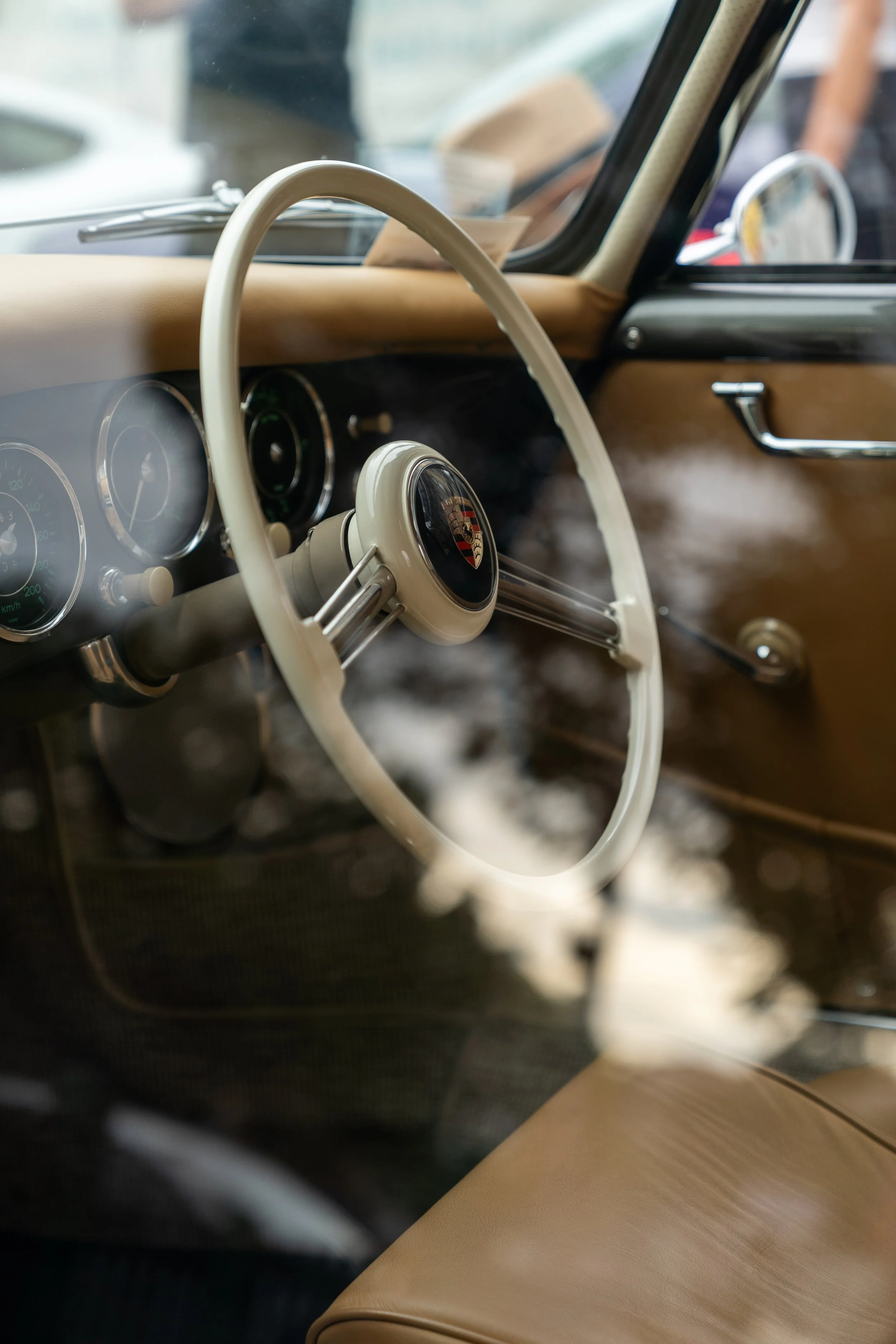 Close-up of the interior of a vintage car, showing a beige steering wheel with a Porsche emblem in the center, a beige dashboard, and a side mirror visible through the window.