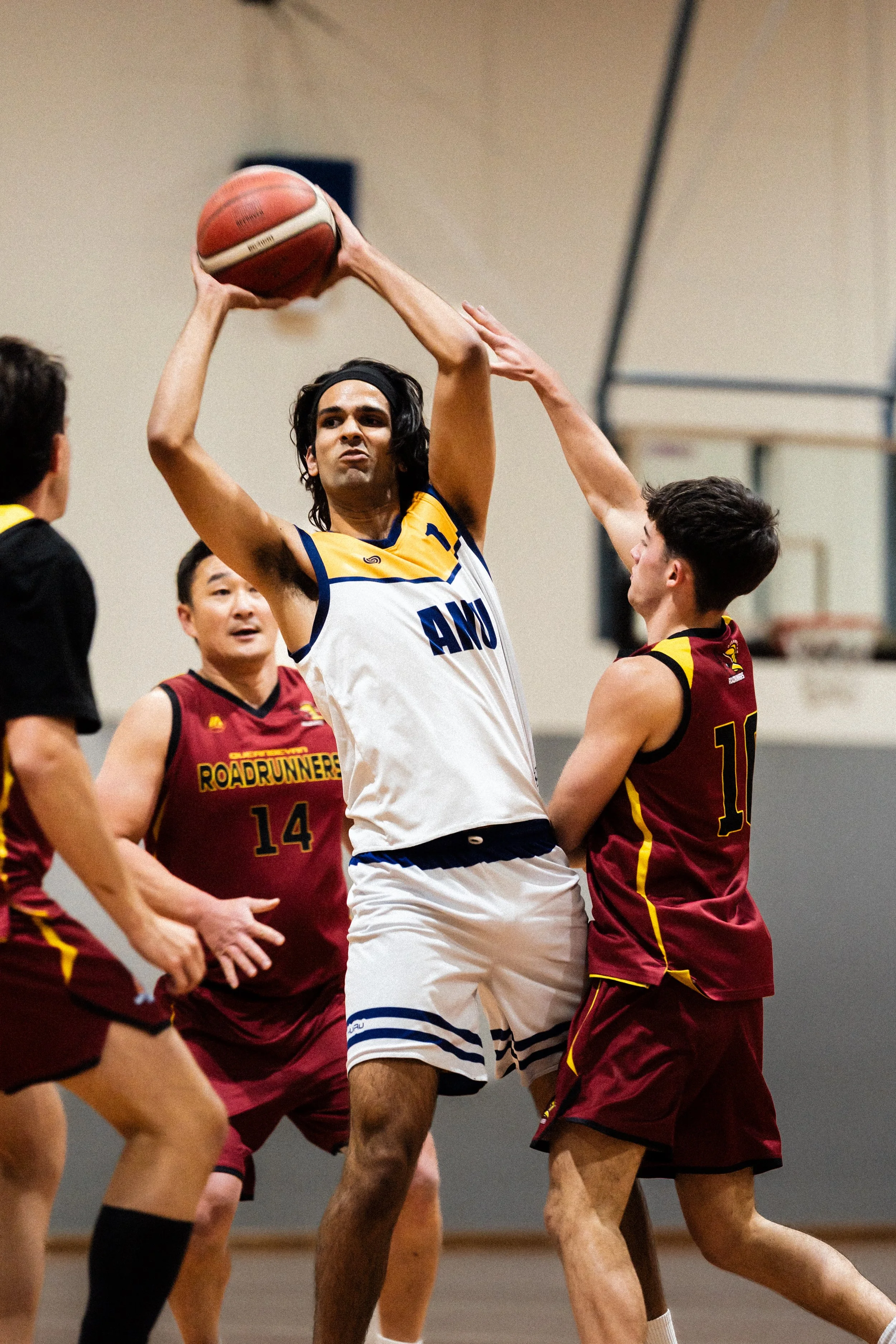A basketball game with players from two teams competing for possession of the ball, with a player in a white and blue uniform trying to shoot or pass while being defended ANU australian national university mens basketball team sports photography