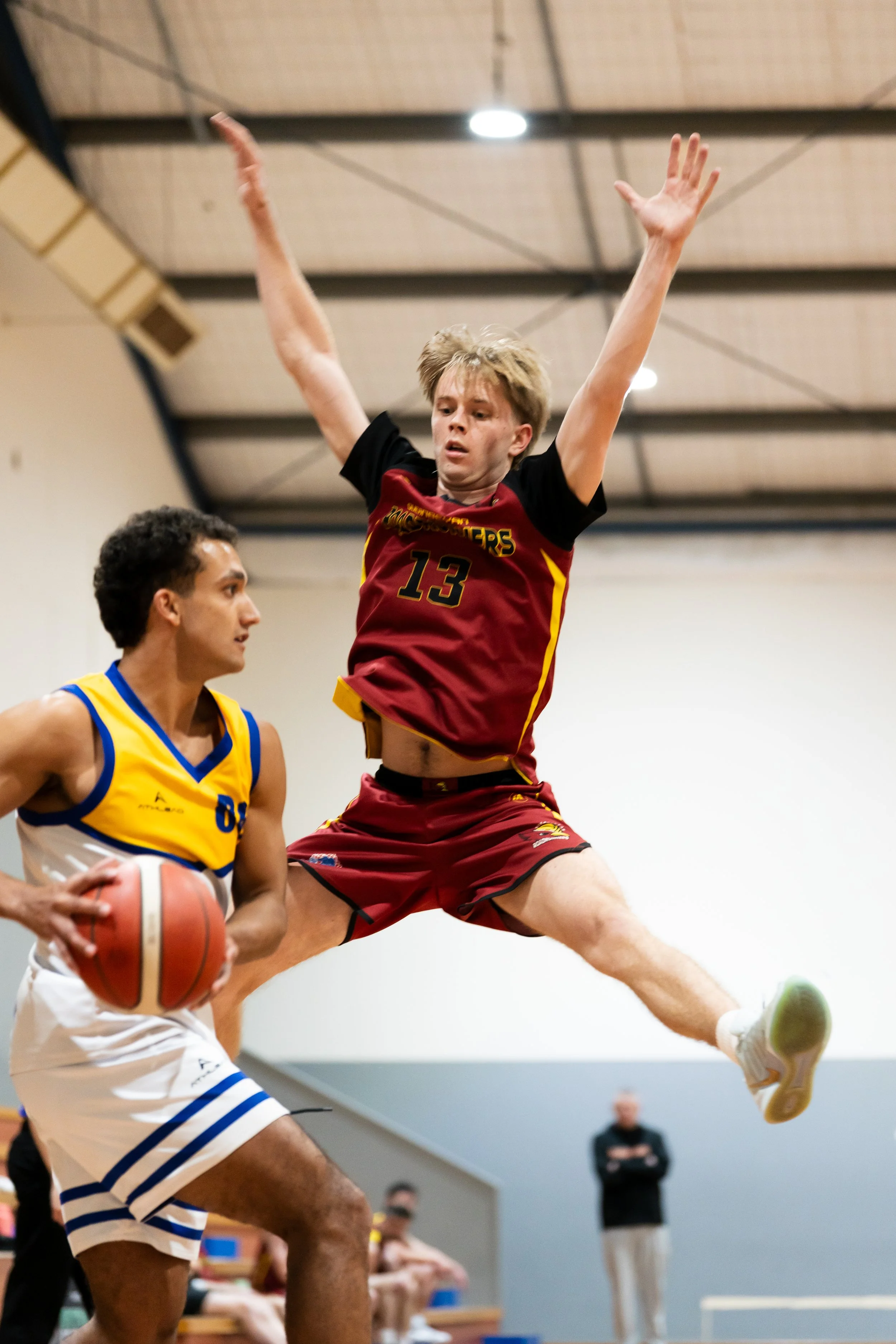 A basketball game in progress, with one player in a maroon uniform jumping to block or shoot, while another player in a yellow and blue uniform holds a basketball, ANU australian national university basketball sports photography