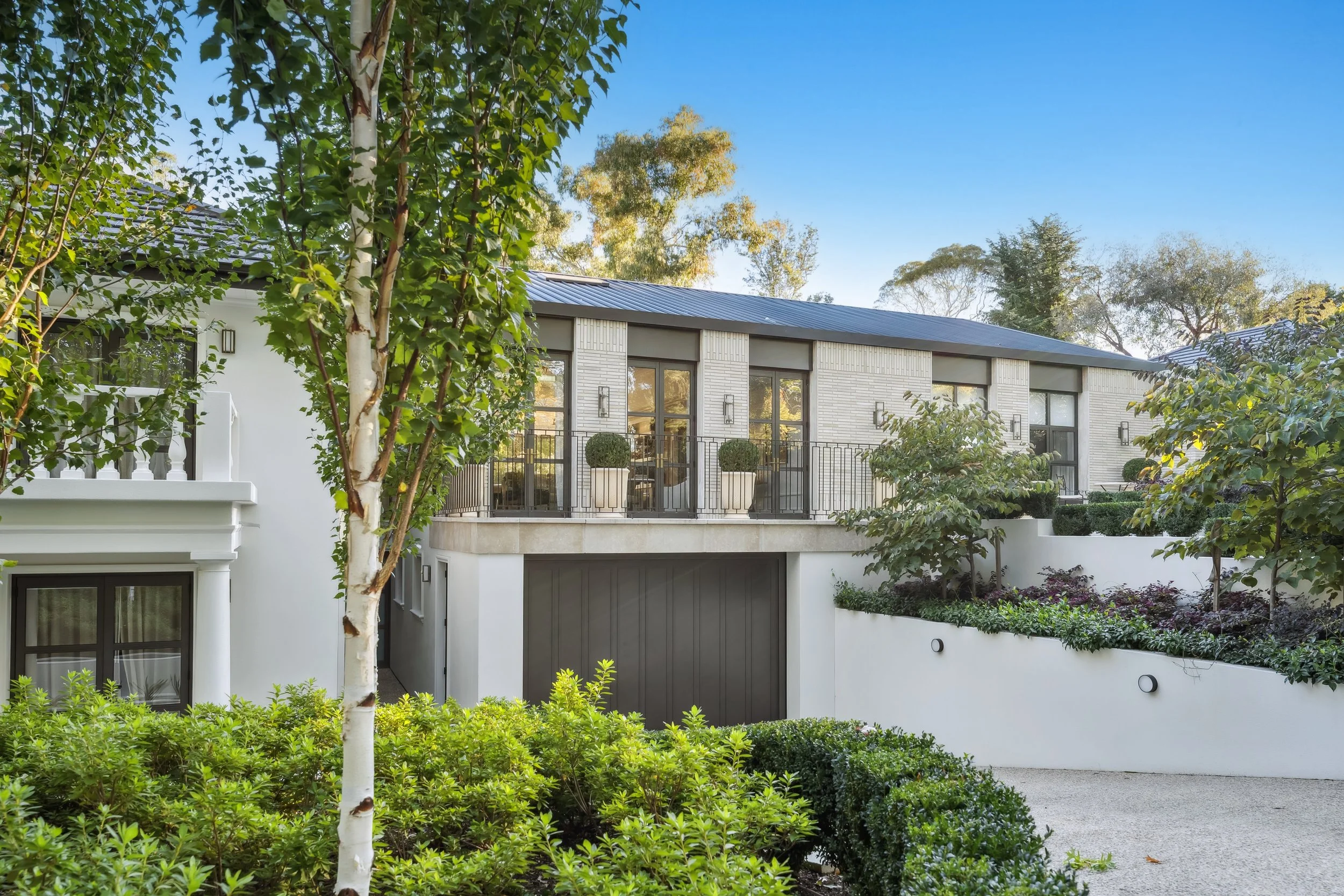 A modern two-story house with large windows and a balcony, surrounded by lush greenery and trees, under a clear blue sky. architecturally designed by thursday architetcure home in red hill editorial photography