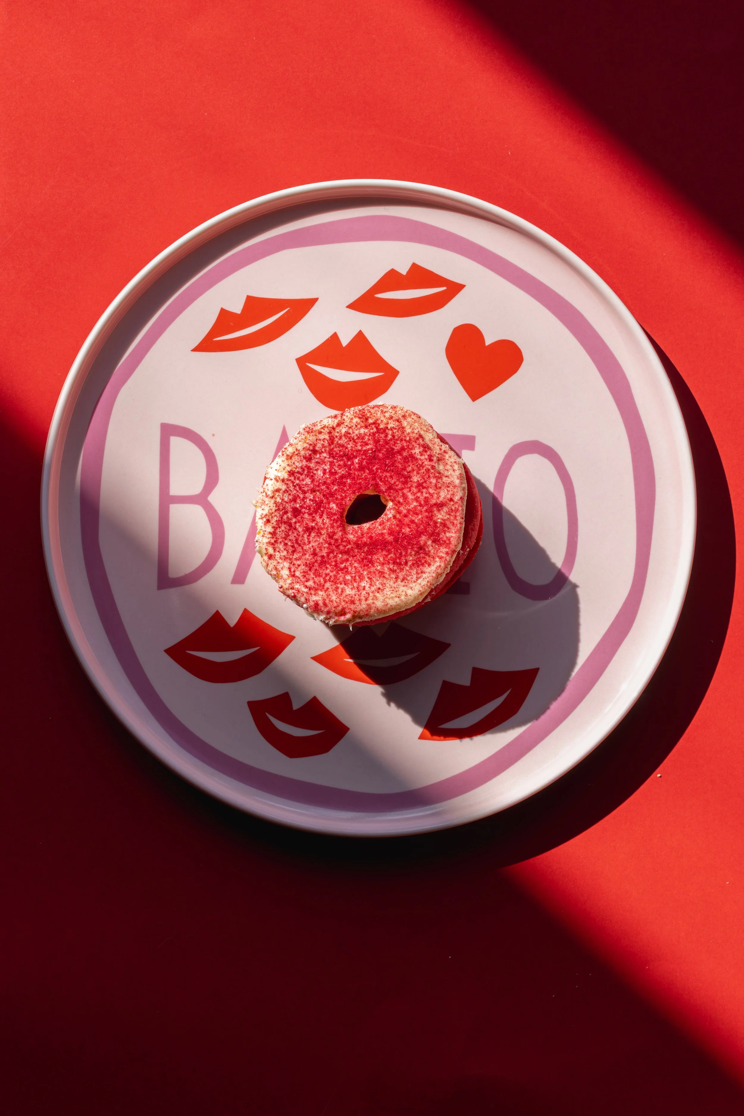 A pink frosted donut with red sprinkles on a plate with lips, kiss marks, and a heart design, set on a red background. round of house pink bacio plate product photography