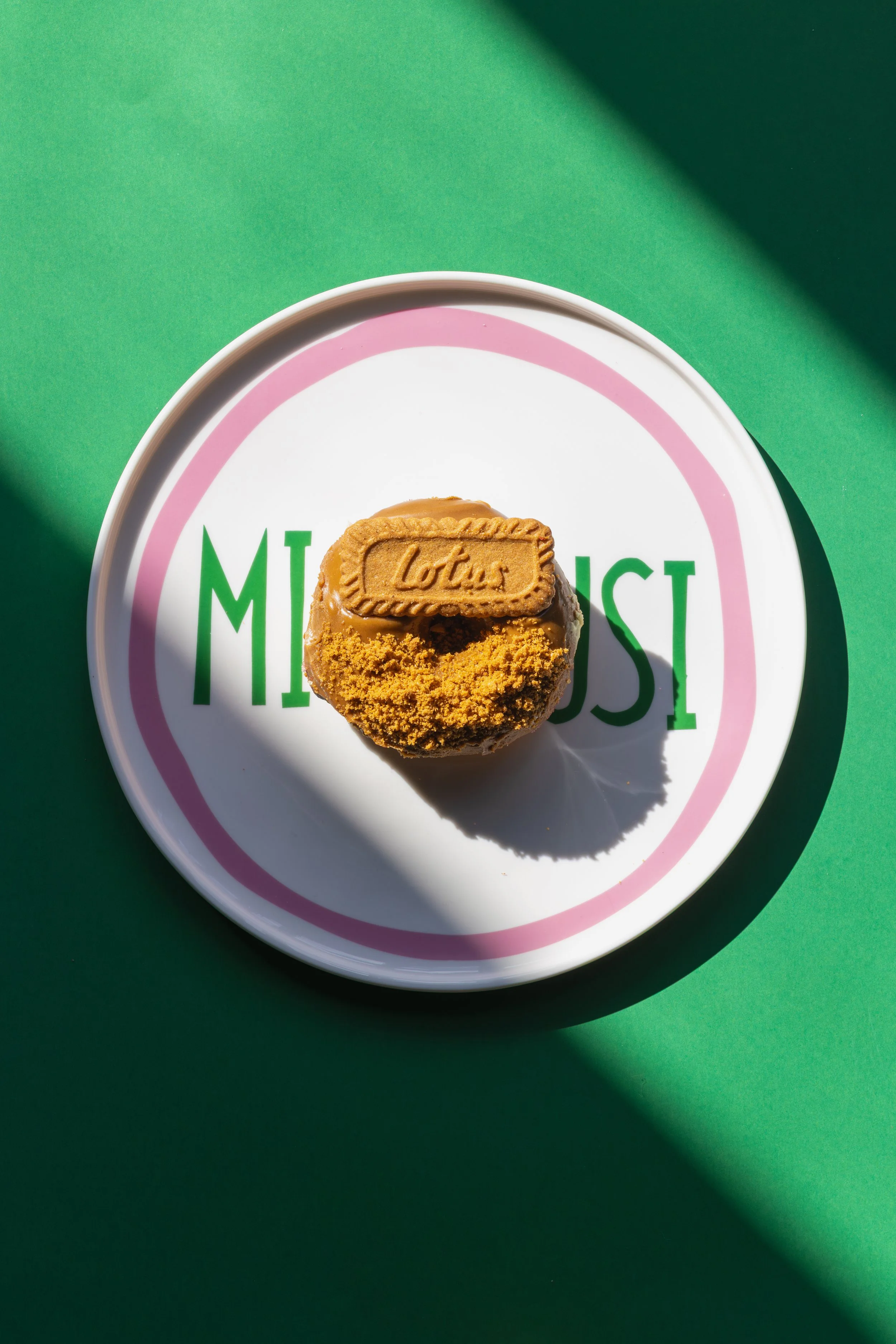 A cookie with a rectangular Lotus Biscoff biscuit on top, served on a white plate with pink and green design, on a green background. round of house plate mi scusi