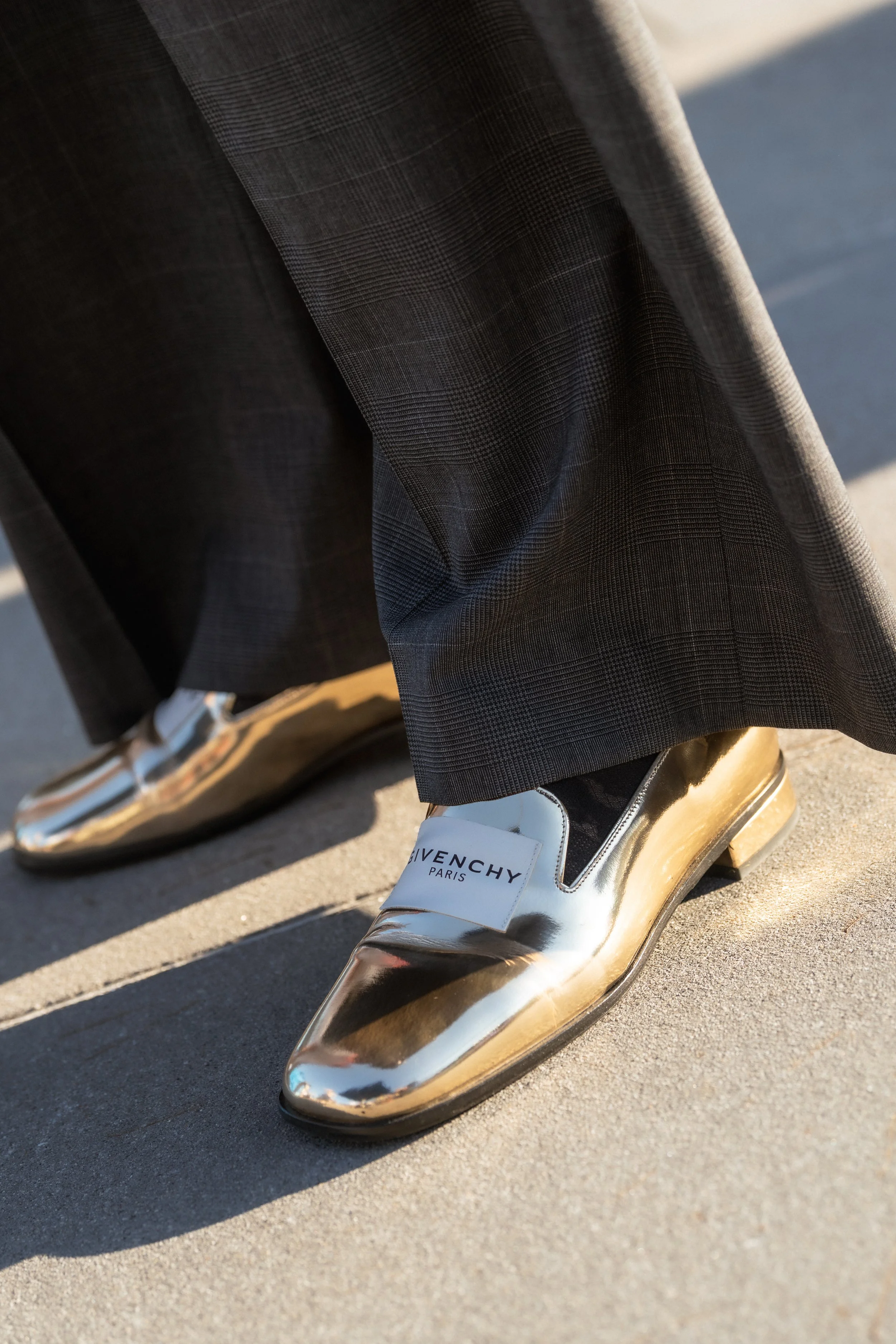 Close-up of a person's legs and feet, wearing black and white Givenchy Paris sneakers with metallic gold heels, dark trousers, and standing on a pavement.