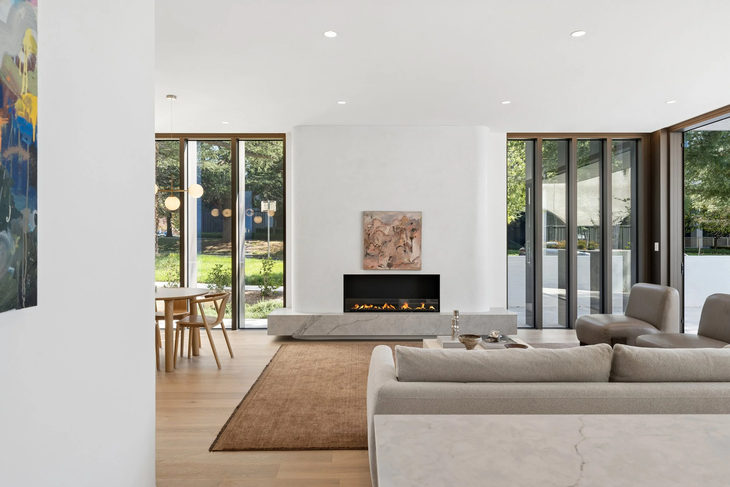 Modern living room with a white ceiling, recessed lighting, a fireplace with a marble surround, and a large window showing outdoor greenery. architectural real estate editorial photography for sotheby's real estate in Forrest canberra
