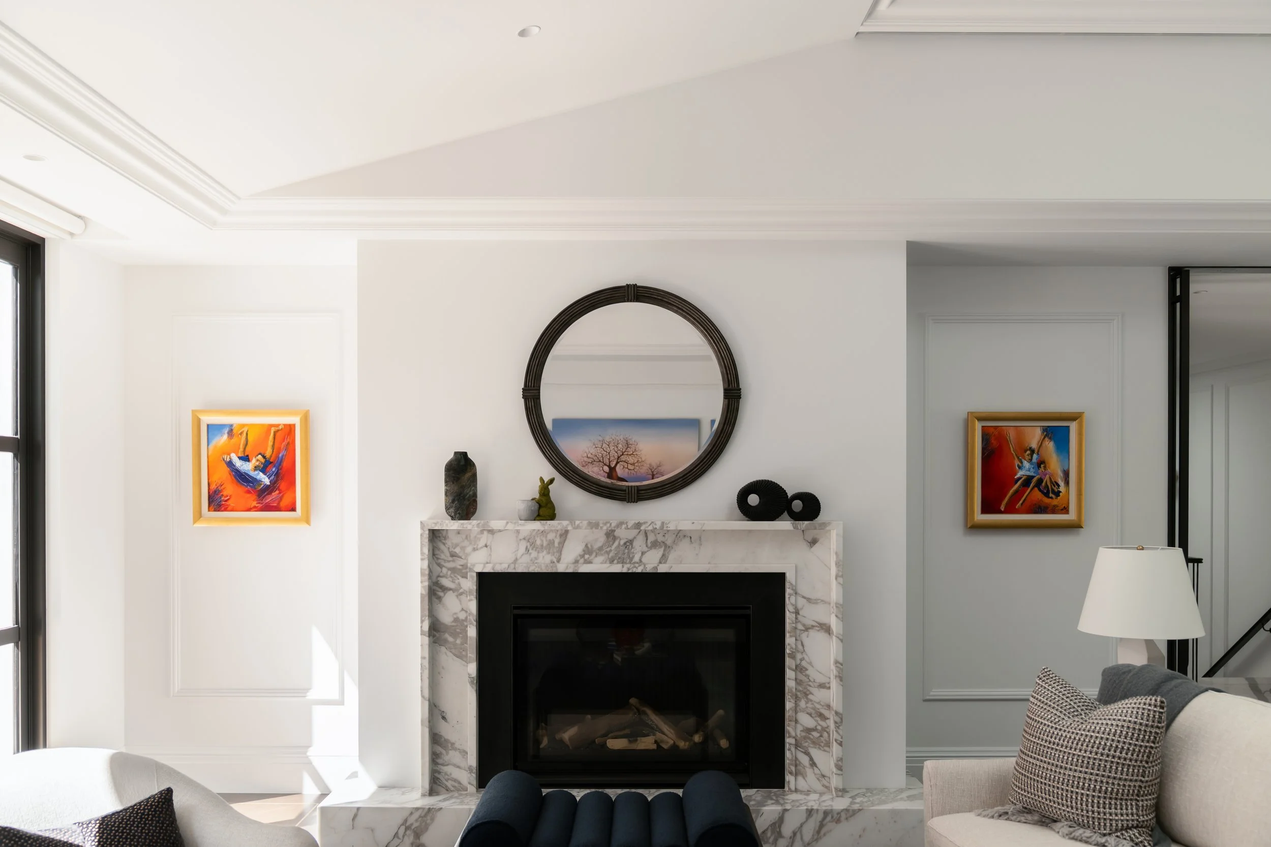 Living room interior with a fireplace, artwork, and decorative items on the mantle. Modern design with white walls, a large mirror, and framed colorful art. architecturally designed interior design by thursday architecture - photography