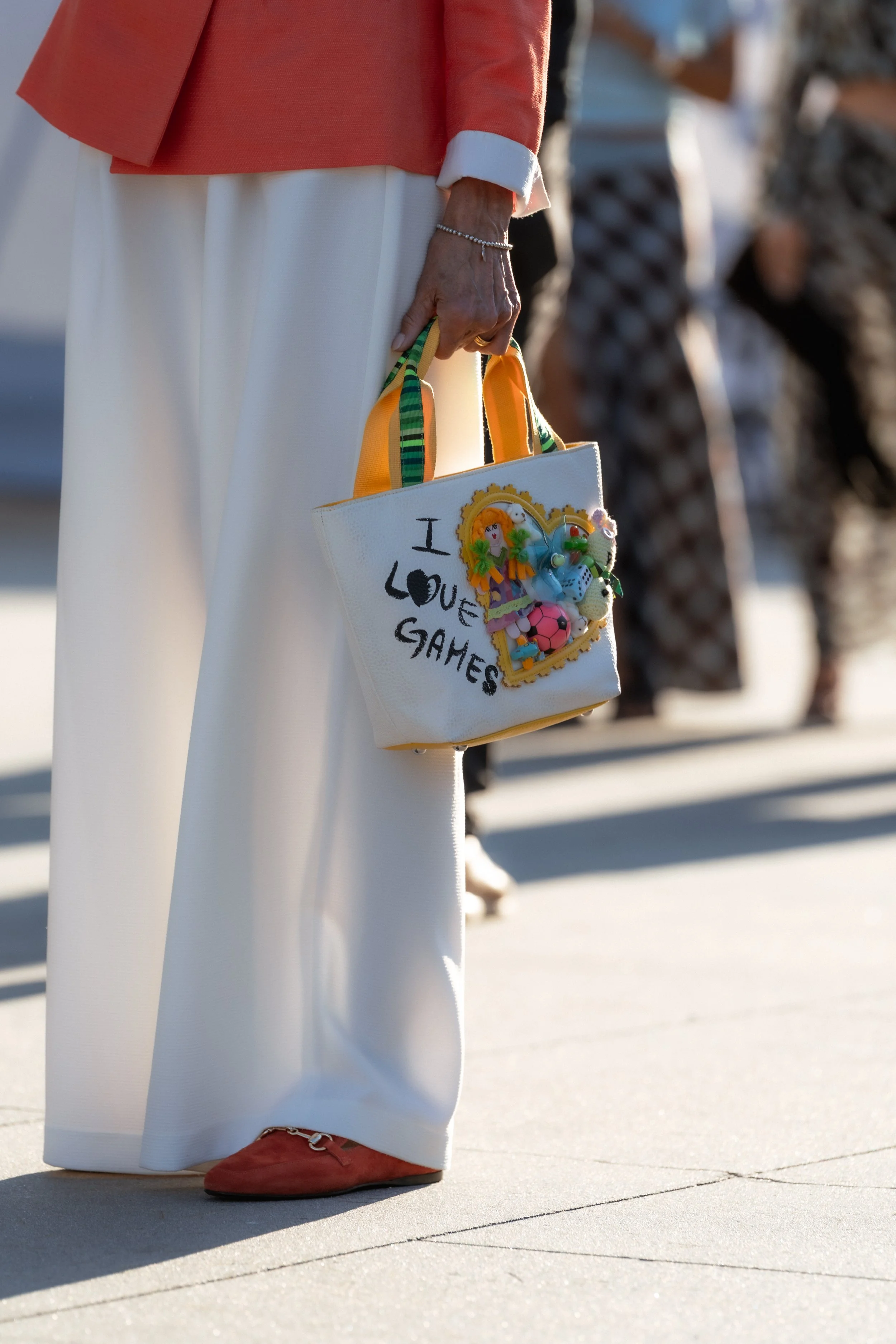 Close-up of a woman holding a small white tote bag with colorful embellishments, wearing white wide-leg pants, a coral jacket with white cuffs, and red shoes with a decorative buckle, standing on a concrete sidewalk.