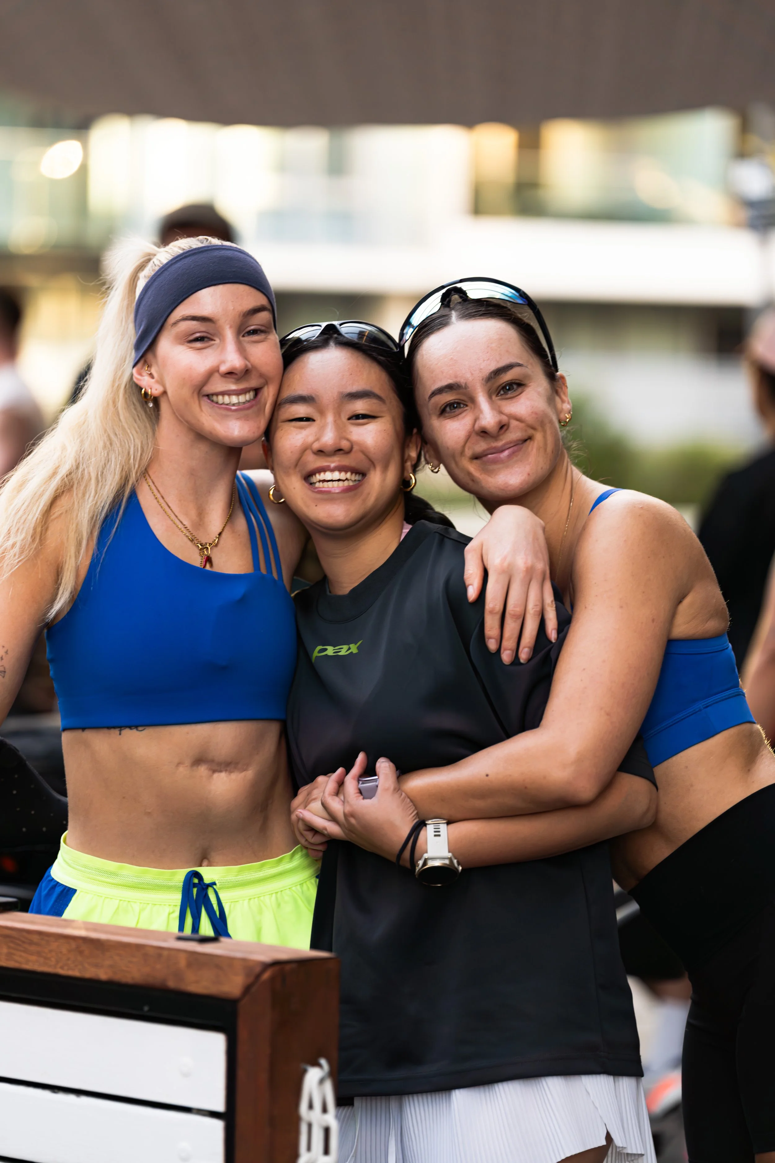 Three smiling women hugging each other outdoors during daytime, wearing athletic clothing. sunday run club canberra PAX LSKD sports photography