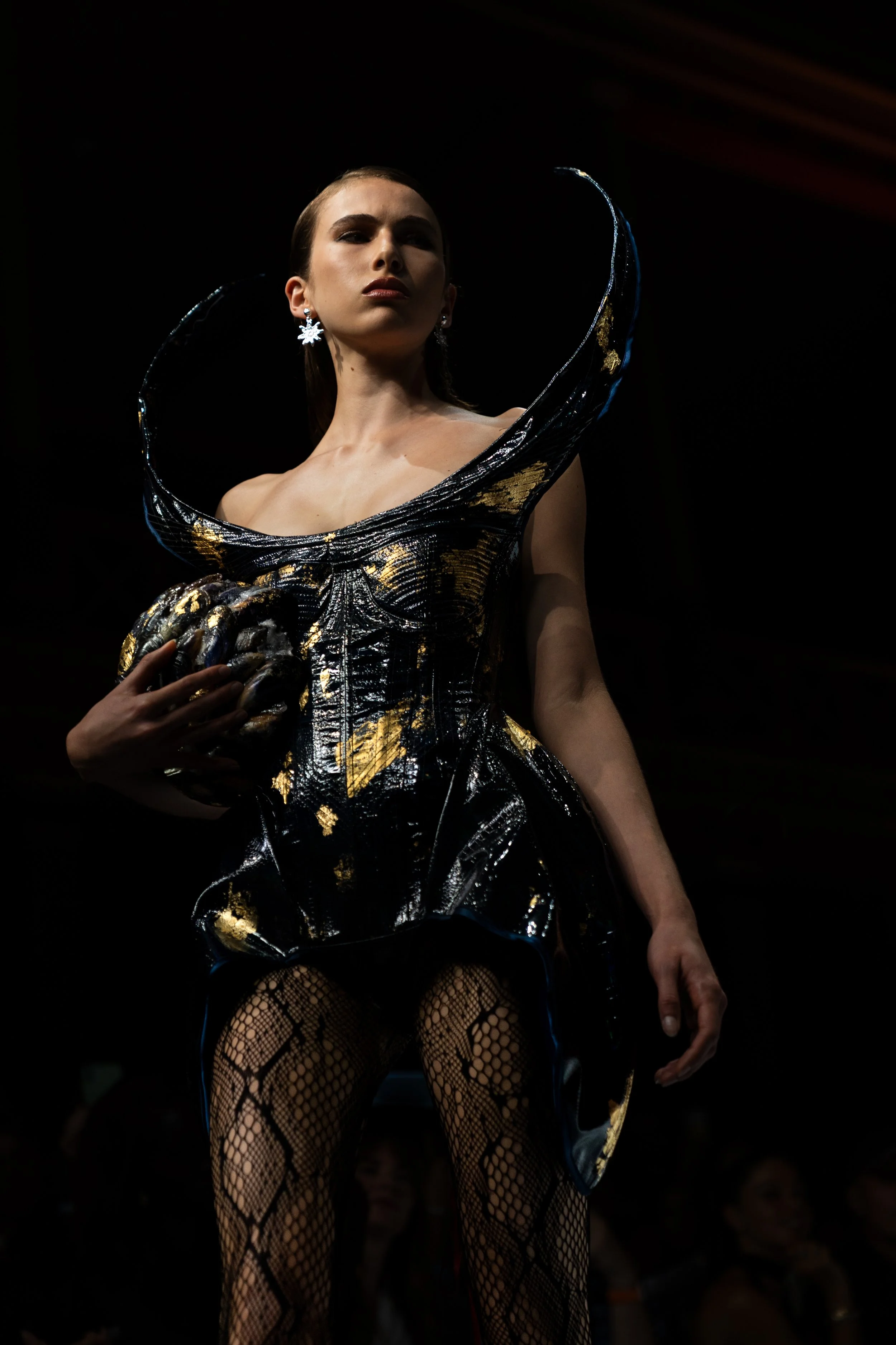 A woman in a metallic, black and gold dress with dramatic shoulder accents, wearing snowflake earrings, fishnet stockings, and holding a textured clutch, poses on a dark runway with a spotlight highlighting her. Melbourne fashion festival fashion