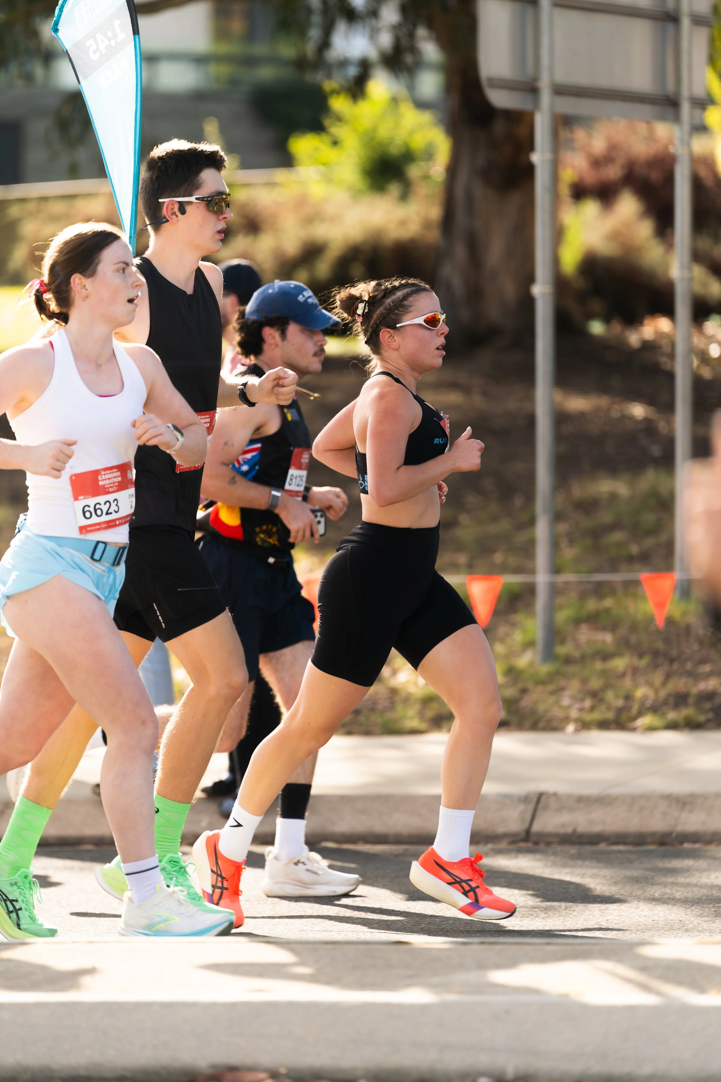 Group of runners participating in a marathon, running on a city street with trees and a sign in the background, wearing athletic clothing and sunglasses. Always in motion sunglasses at canberra marathon sports photography