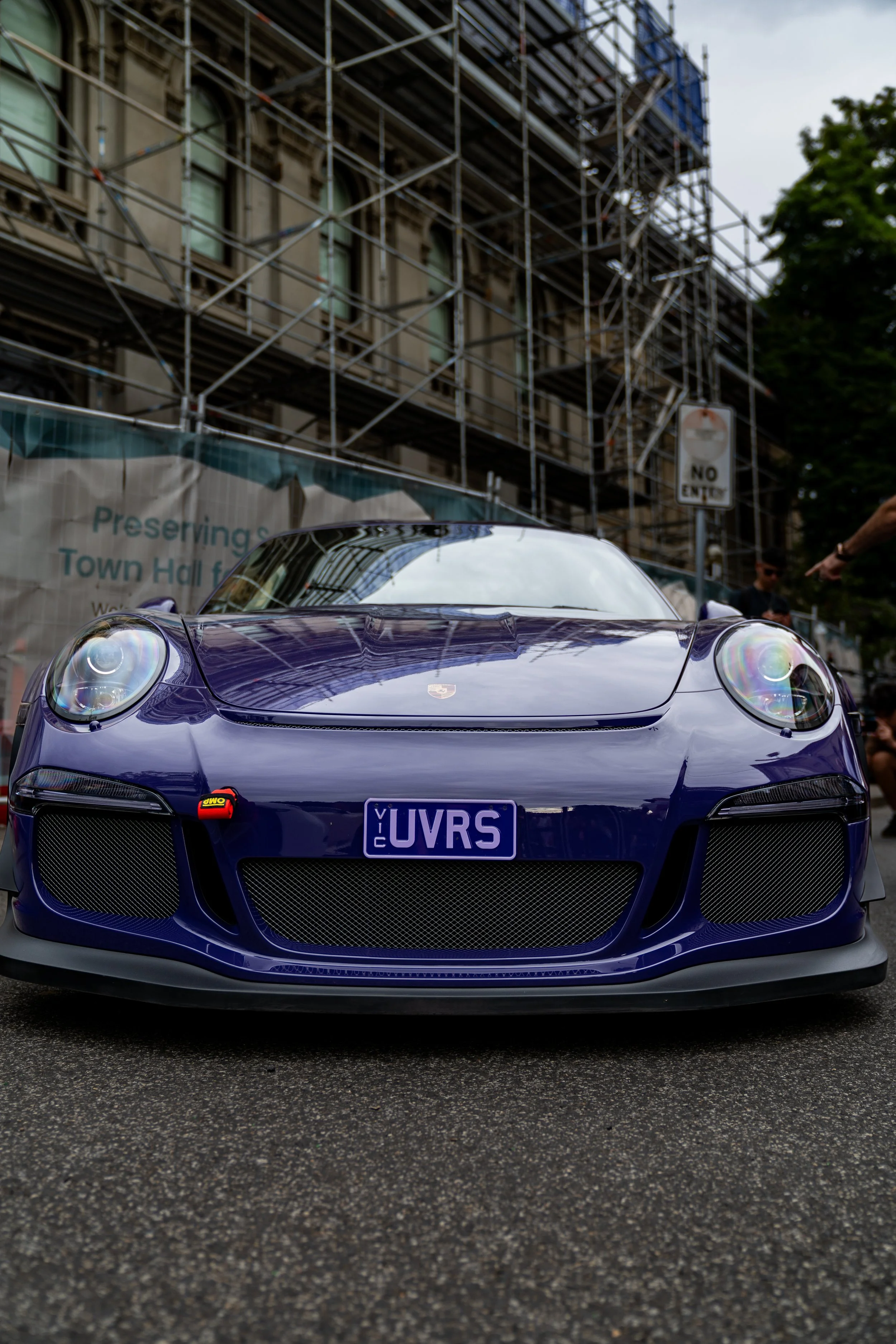 A purple sports car with a California license plate "UVRS" parked on a street, with scaffolding and buildings in the background. Porsche 911 GT3 RS purple custom wrap 