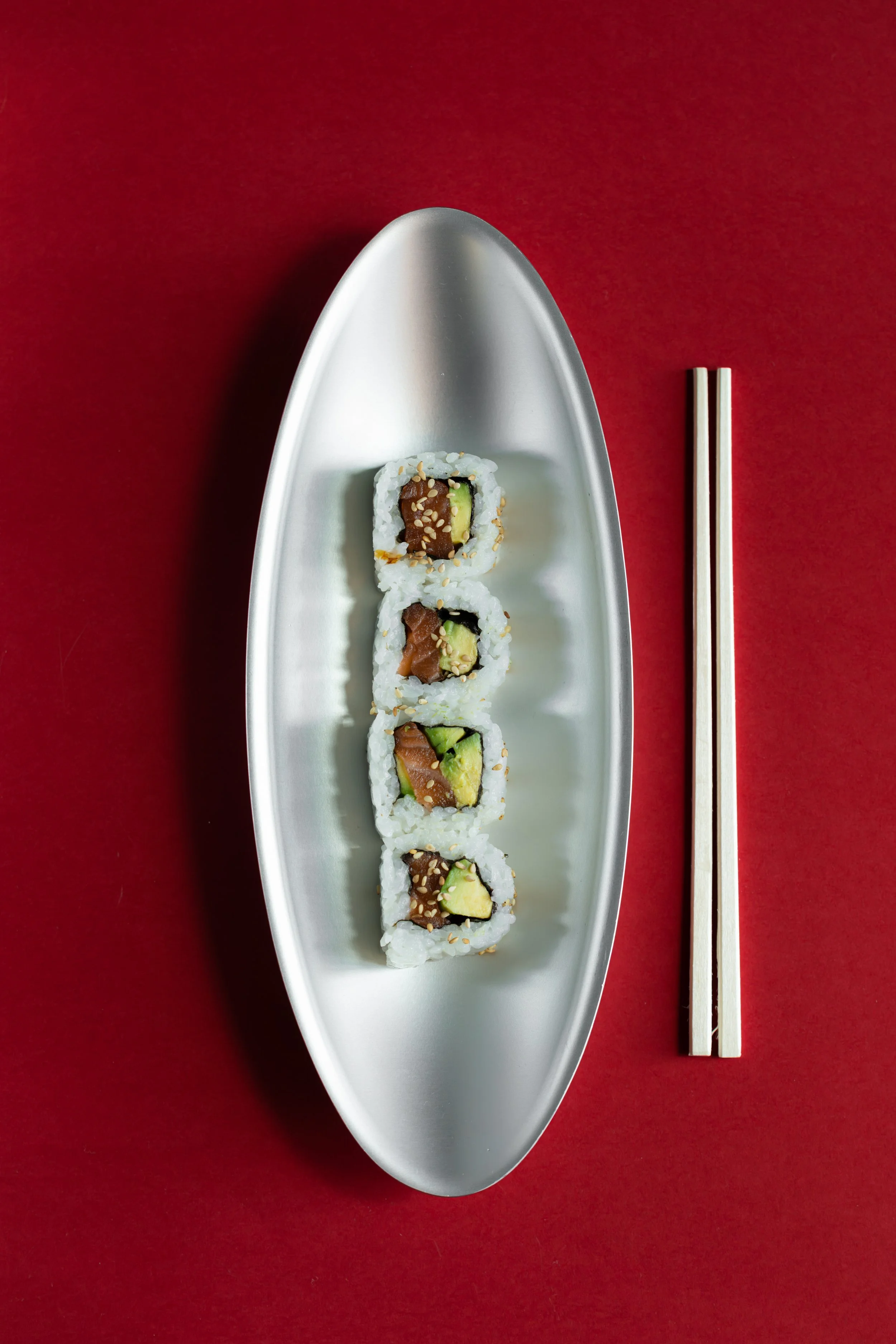 Four sushi rolls with avocado, salmon, and sesame seeds on a silver oval plate, with wooden chopsticks on a red background. fink dish product photography