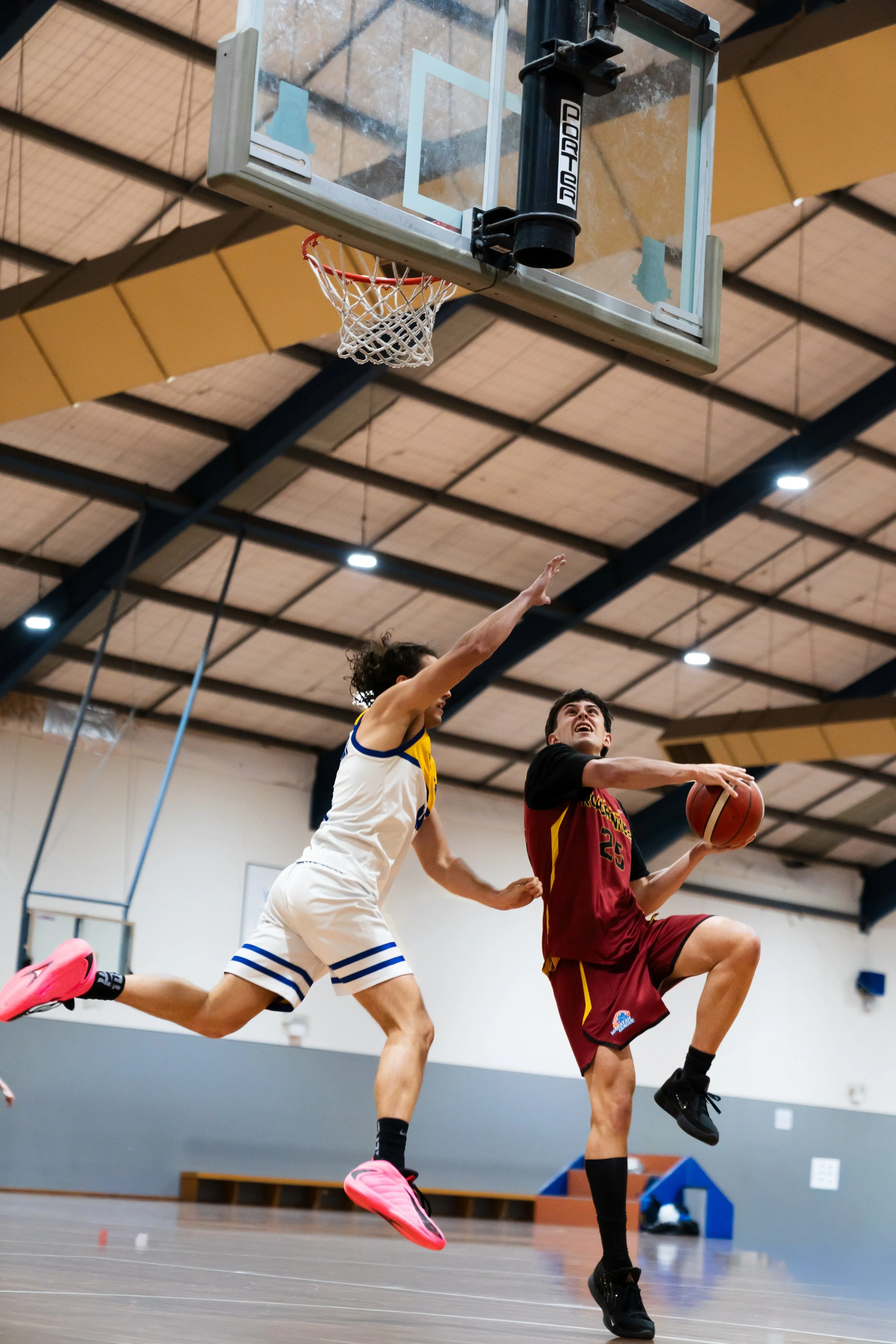 Two basketball players competing for the ball during a game in an indoor gymnasium with a basketball hoop visible in the background. ANU australian national university basketball sports photography