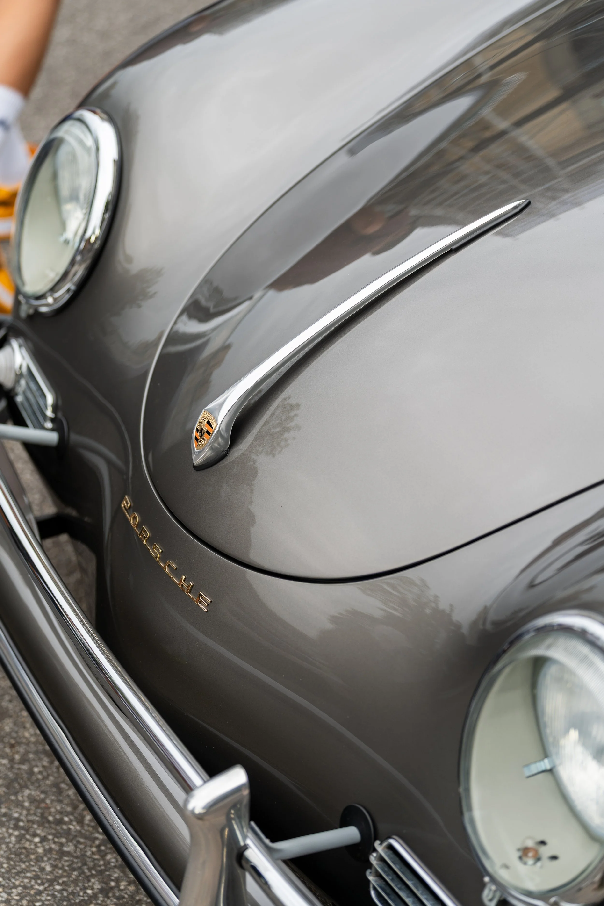 Close-up of a vintage Porsche car's front, showcasing the headlight, part of the hood, and the Porsche logo badge in gold on a gray paint finish, with reflections on the surface.