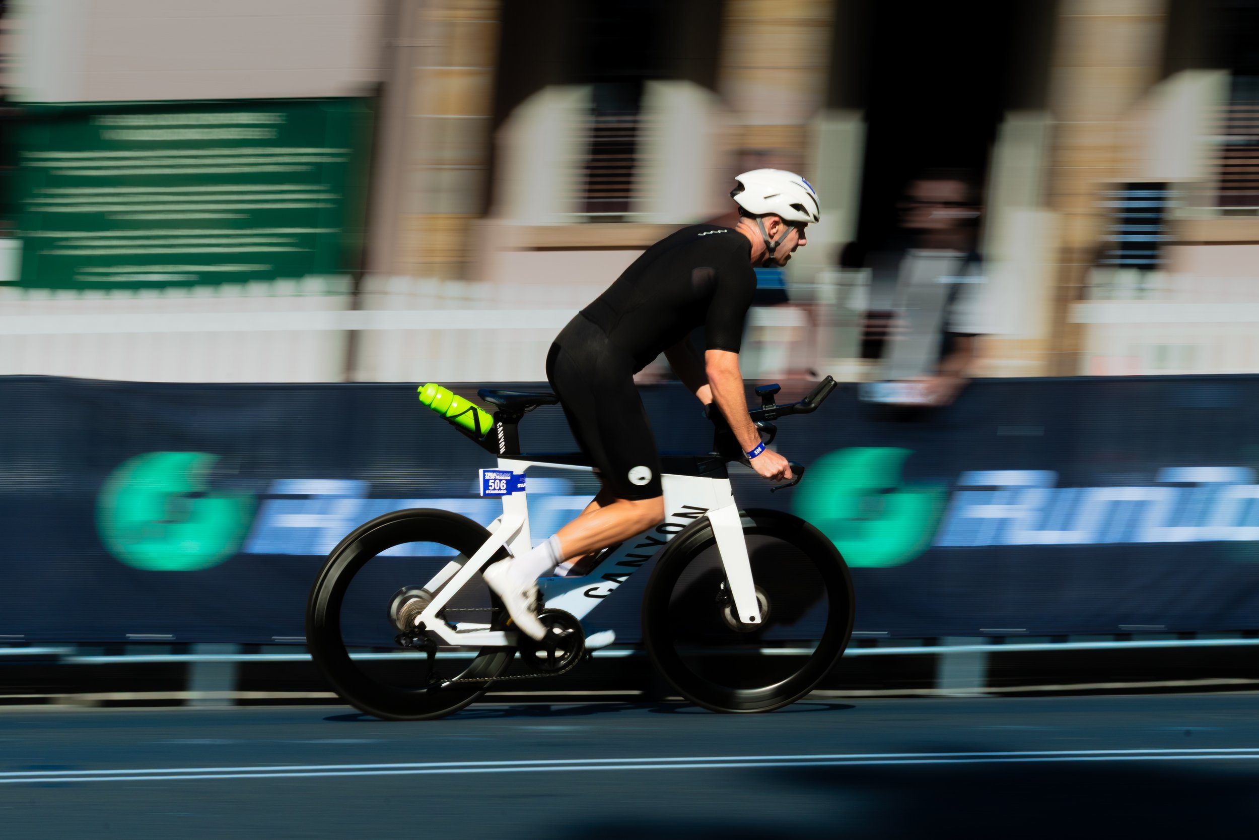 A male cyclist wearing a white helmet and black clothing riding a white bicycle on a road with blurred background. canyon triathlon cycling photography wollongong