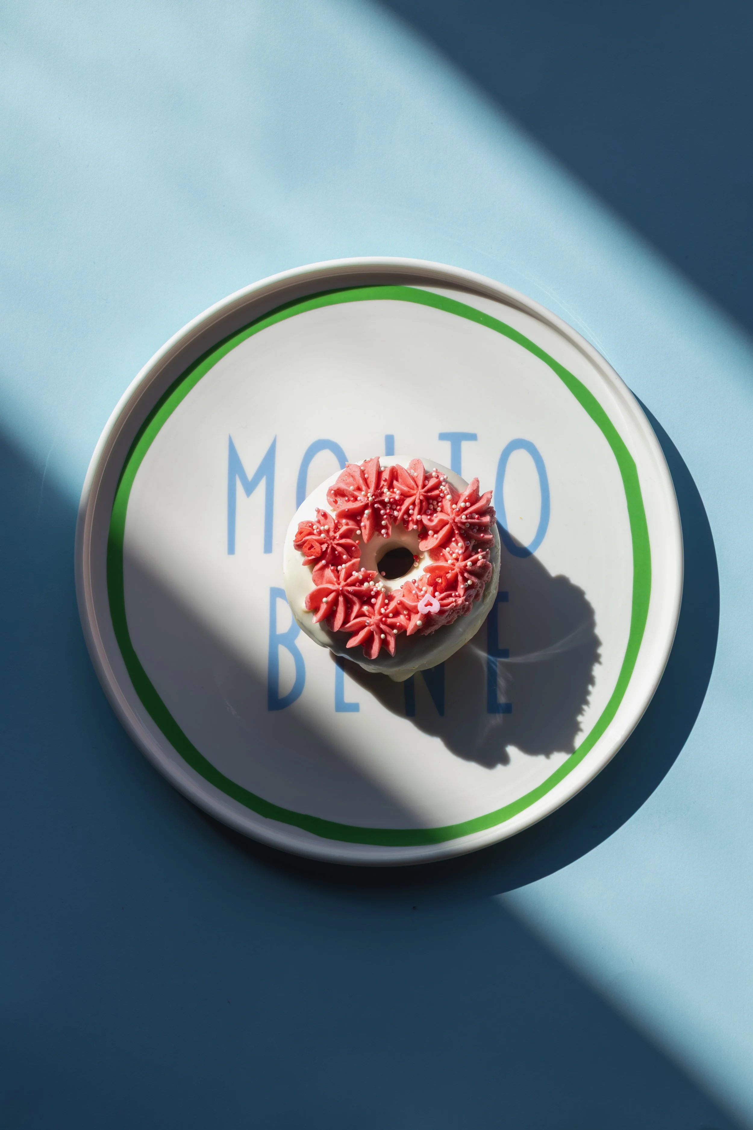 A donut with pink frosting and sprinkles on a round white plate with green border, placed on a blue background with light shadow. round of house molto bene plate product photography 