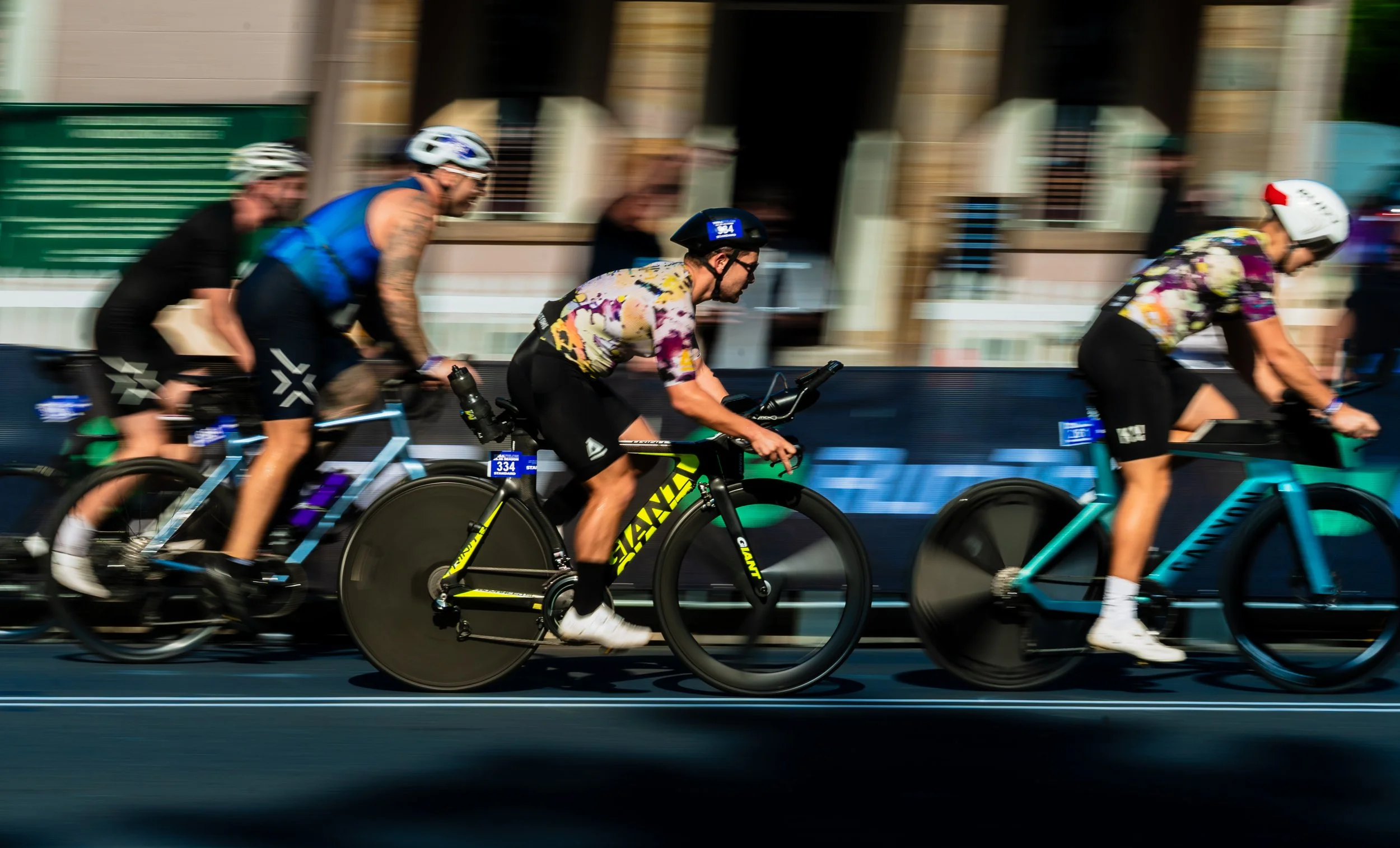 Group of four cyclists racing on a road, with motion blur indicating speed, wearing helmets and colorful athletic clothing. Giant road bike at wollongong triathlon 2026 sports photography