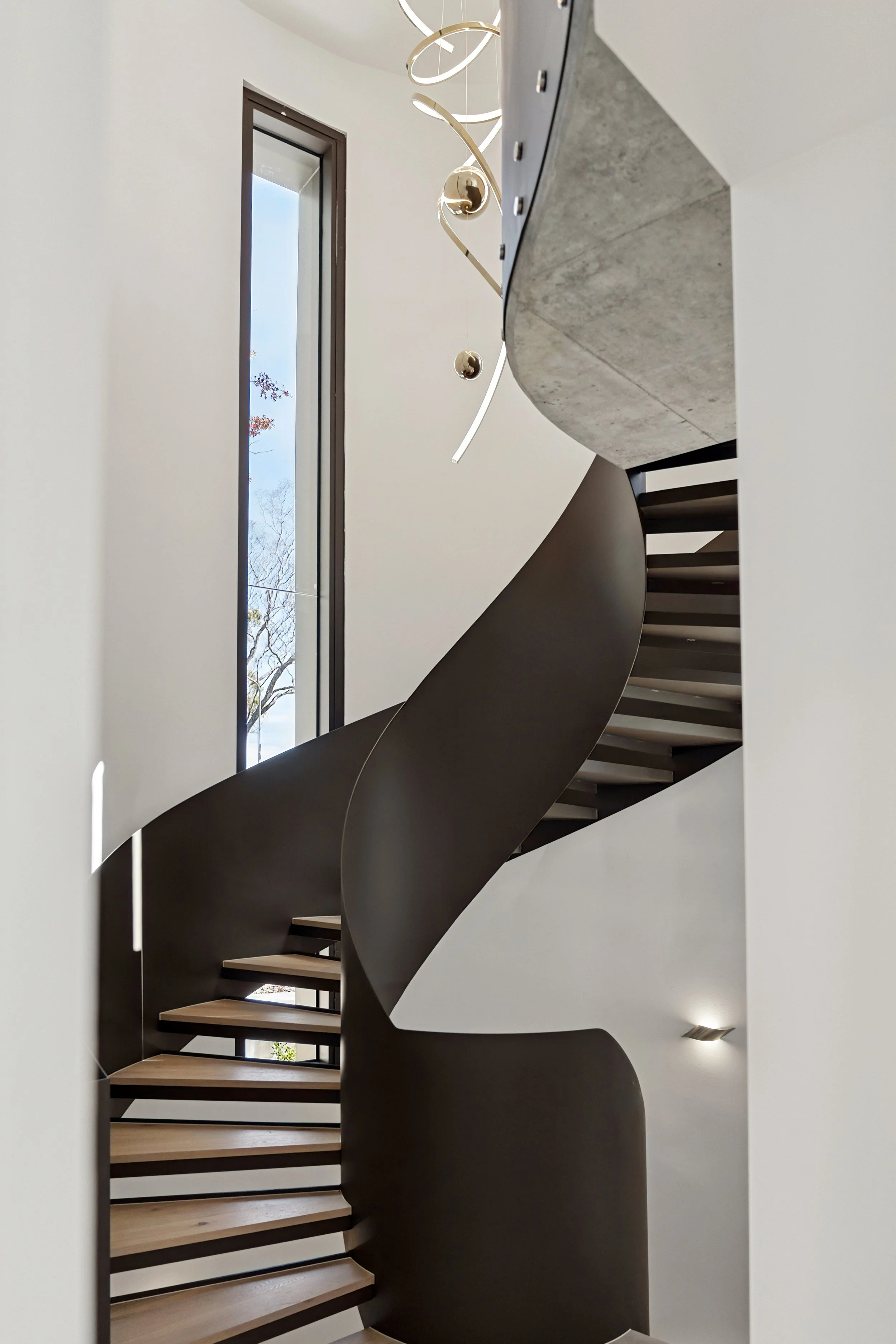 Modern spiral staircase with wooden steps and black railing inside a building with white walls and a tall vertical window, allowing natural light to enter. Architecturally designed home in Forrest by Thursday Architecture 