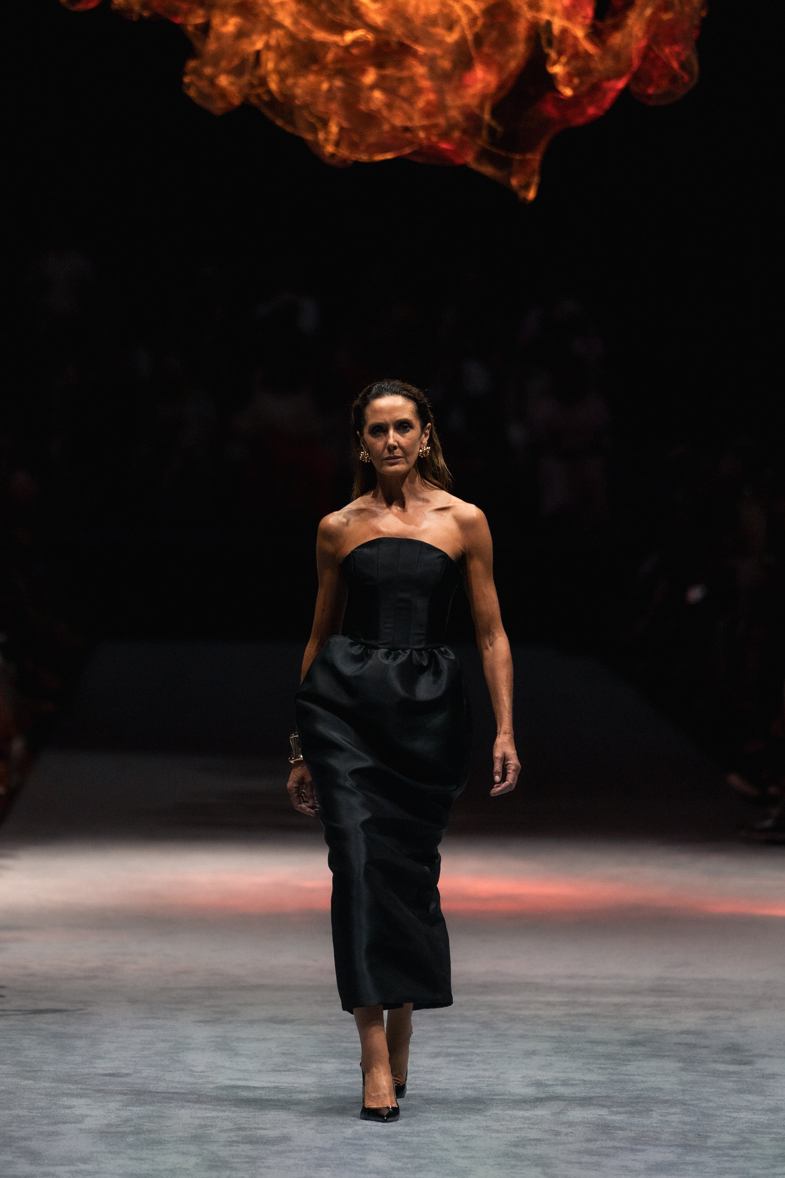 A woman walking on a runway during a fashion show, wearing a strapless black dress and gold earrings, with a fiery orange fabric floating above her head. melbourne fashion festival effie kats runway show