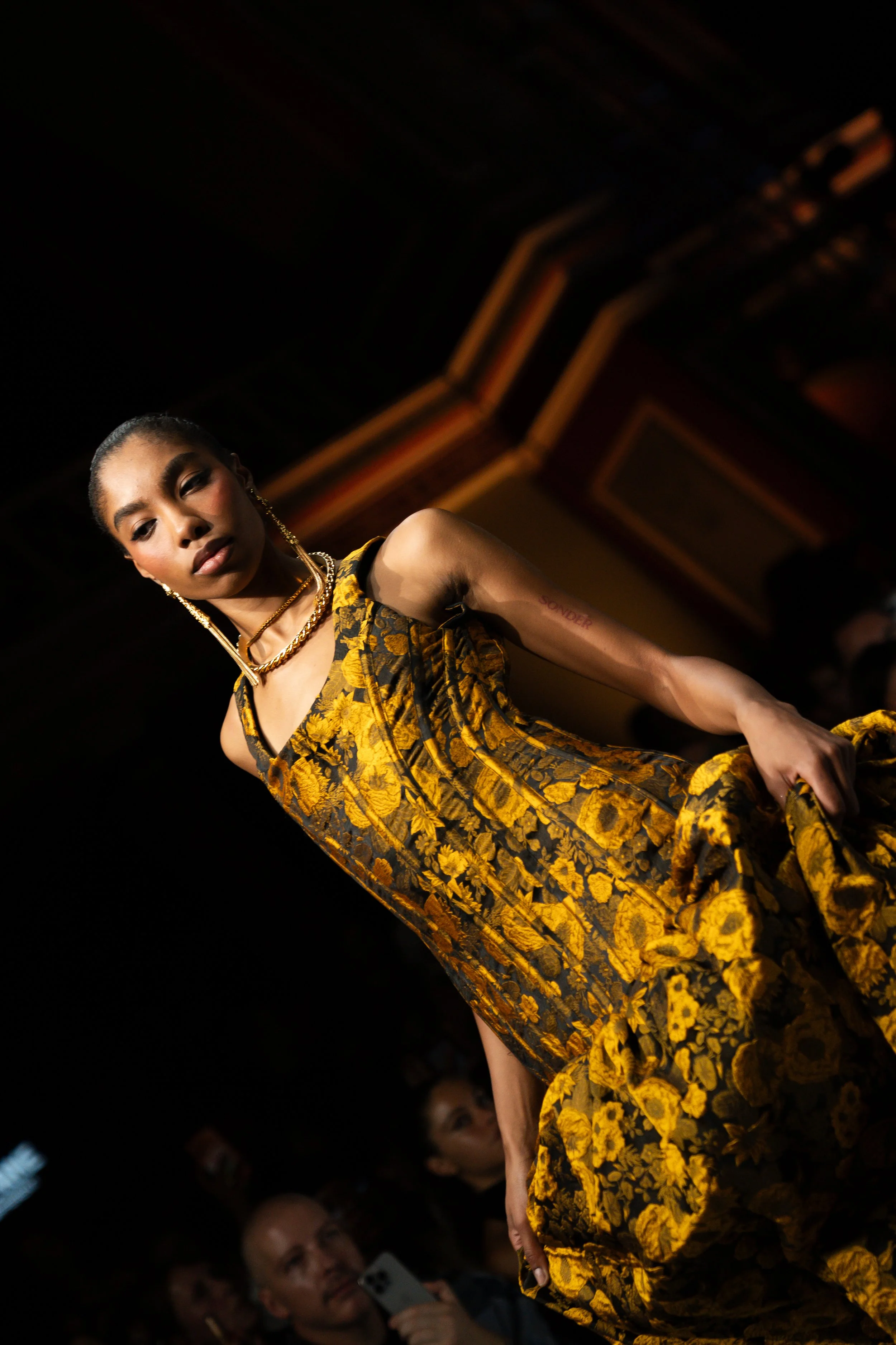 A woman wearing a yellow and black floral dress walking on stage at a fashion show, with an audience watching. melbourne fashion festival Jarrod reid runway show 