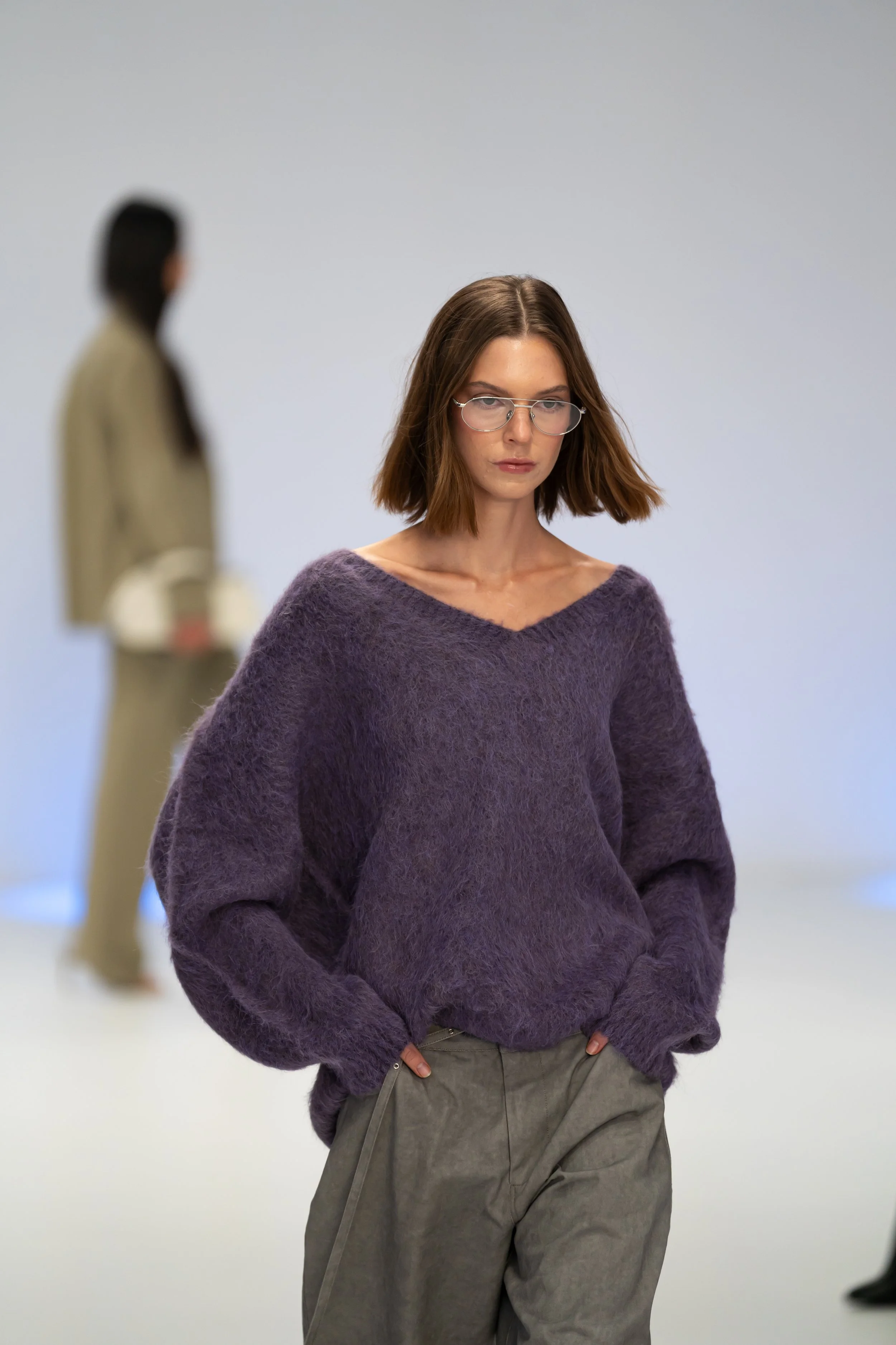 A woman with shoulder-length brown hair wearing glasses, a purple fuzzy sweater, and gray pants stands with hands in pockets. In the background,  Australian Fashion Week 2026 Bec and Bridge, Aje Studio runway Photography