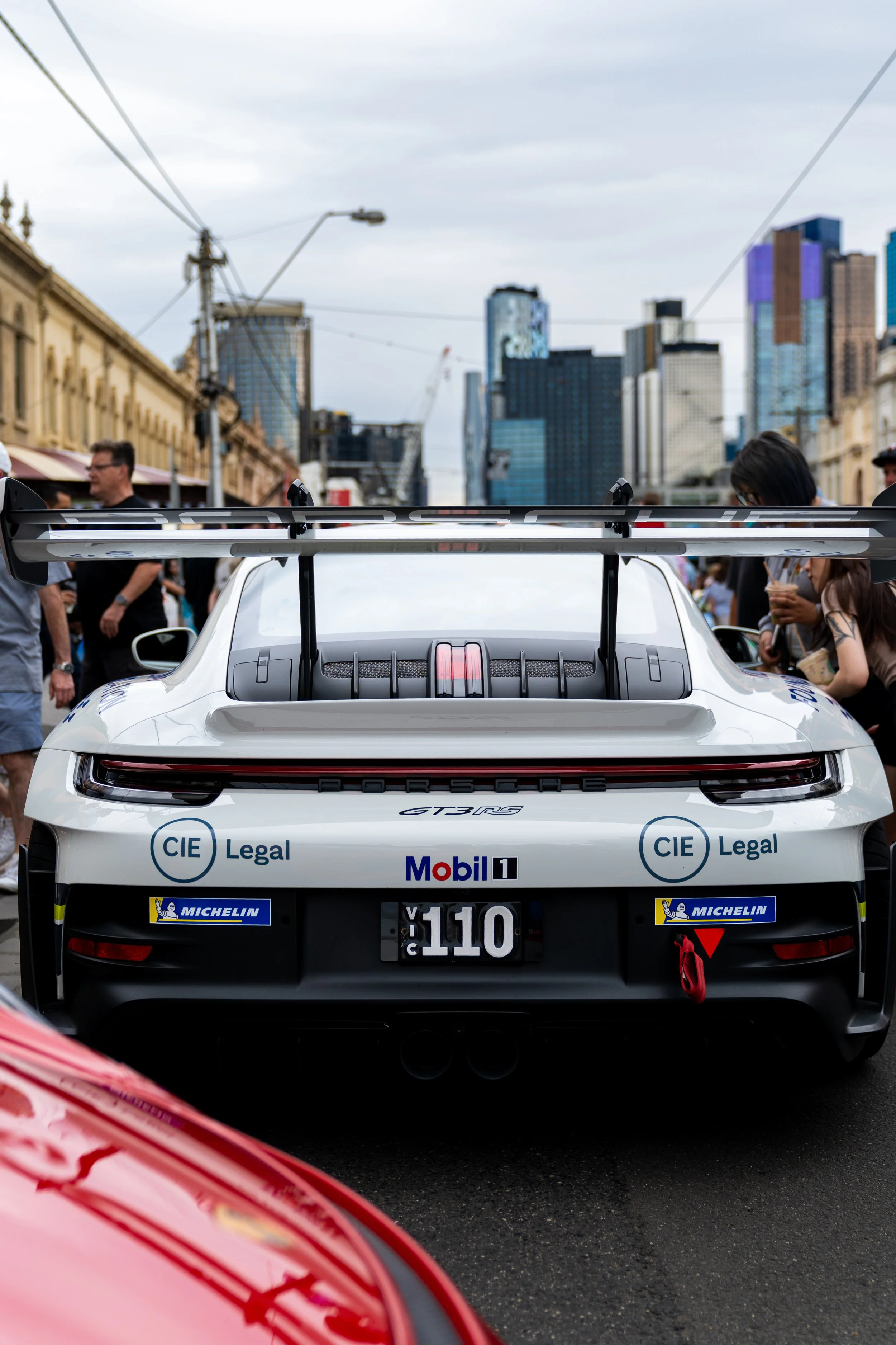 Rear view of a white Porsche race car with sponsor stickers, parked among a crowd in an urban setting with tall buildings in the background. porsche 911 gt3 rs Carrera cup race car heritage VIC number plate