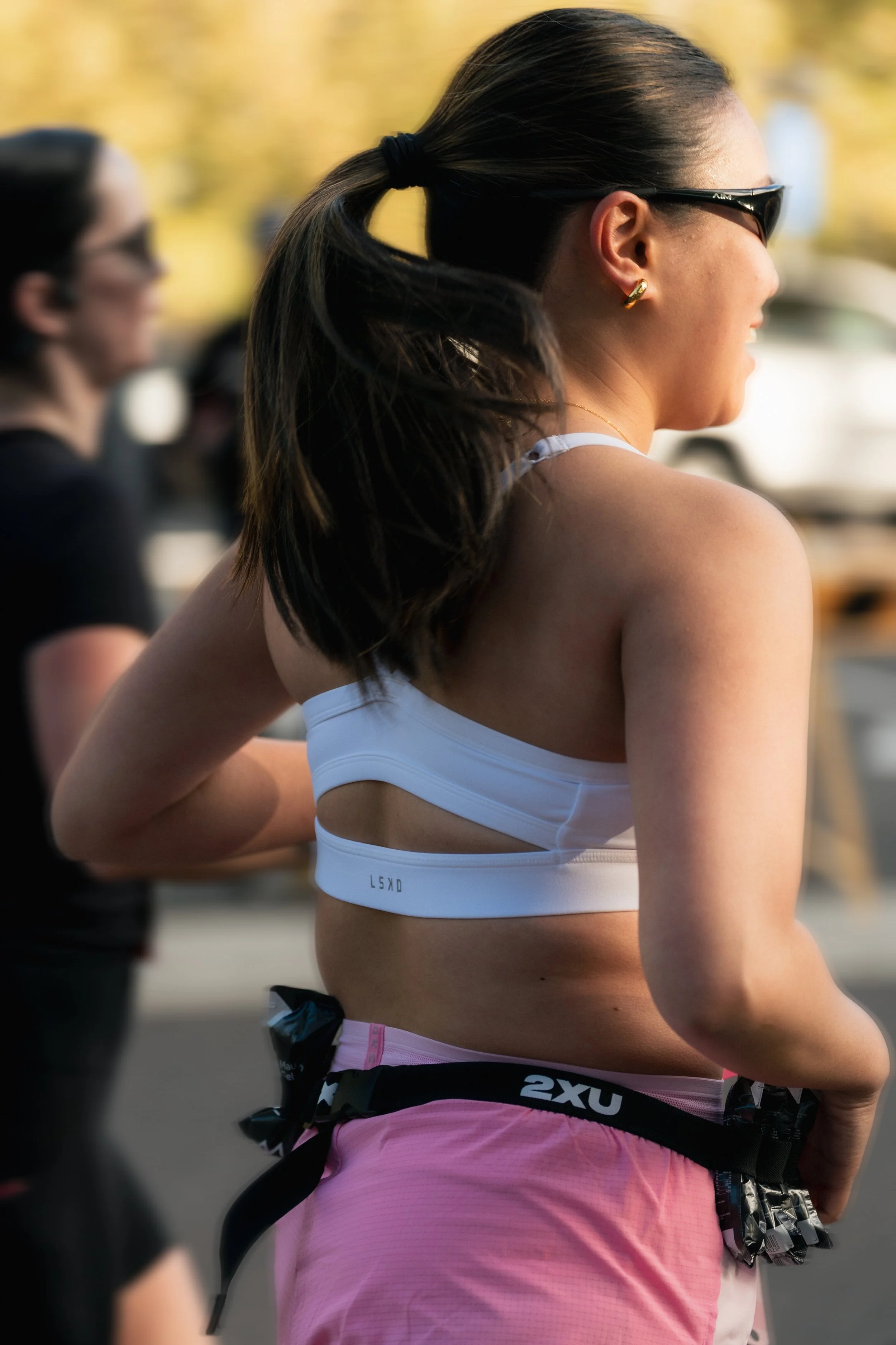 A woman with long dark hair in a ponytail, wearing sunglasses, a white sports bra, and pink athletic shorts, participating in an outdoor running event. LSKD running at Canberra Marathon 2026 sports photography