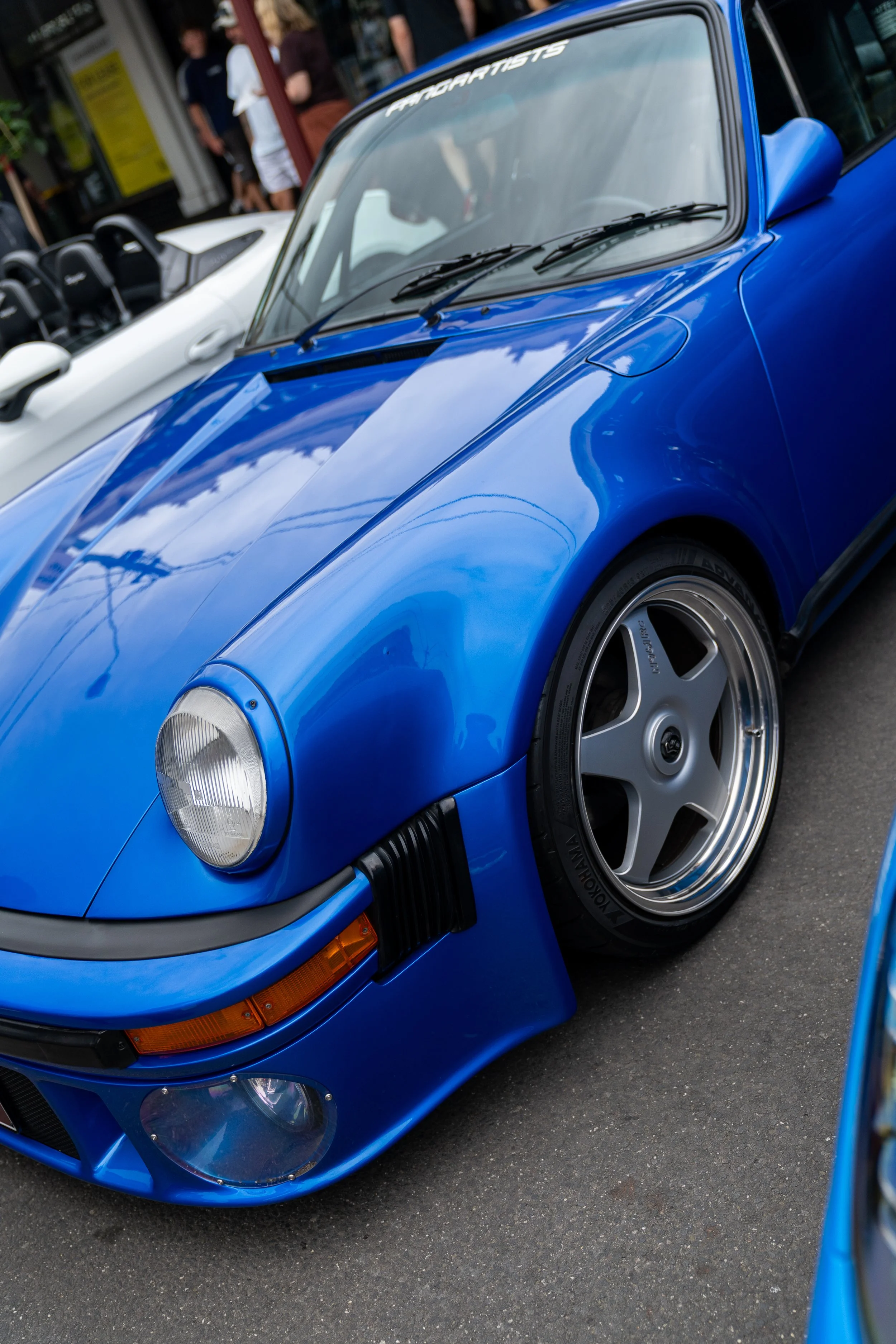 Close-up of a classic blue Porsche sports car parked on the street, with a white car visible in the background and people standing nearby. custom porsche 911 930 turbo blue wrap 