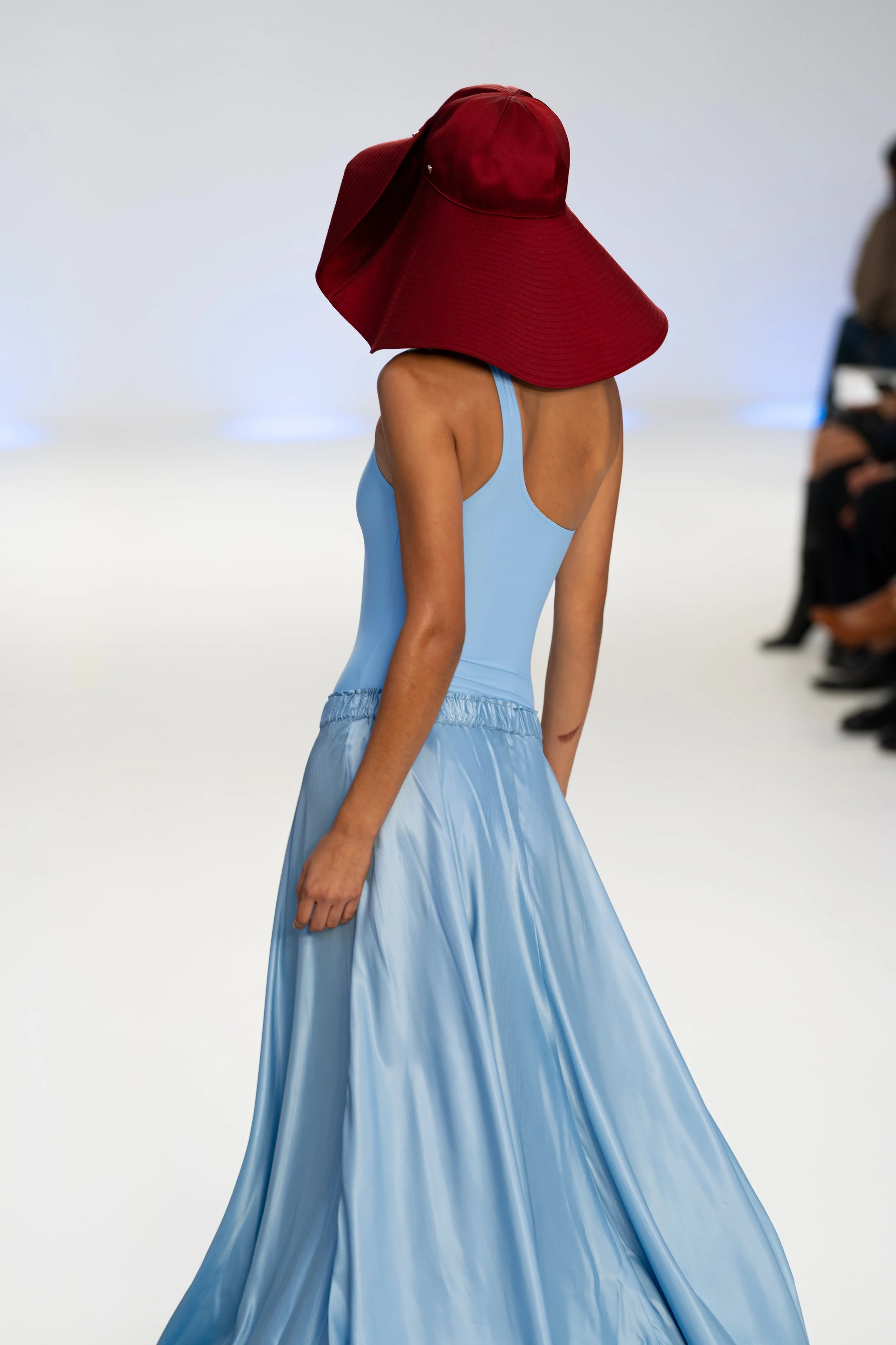 Model walking on runway wearing a light blue dress with large red hat that extends over her shoulders, during fashion show. Australian Fashion Week 2026 the edit runway photography