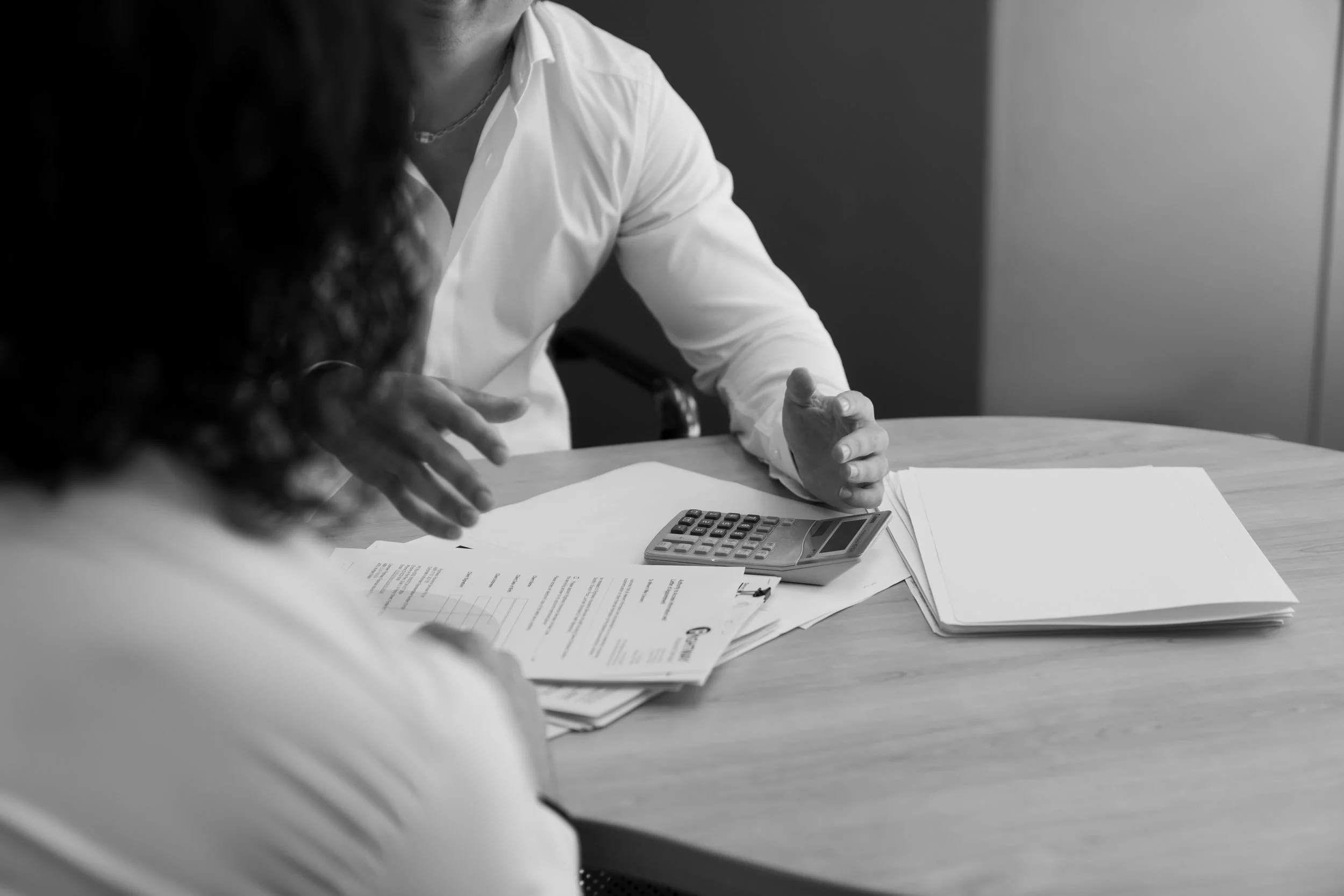 Black and white photo showing two people in a serious business discussion at a table covered with documents, a calculator, and a pen, one person is mostly out of frame while the other, wearing a white shirt, is visible from the chest down with hands 