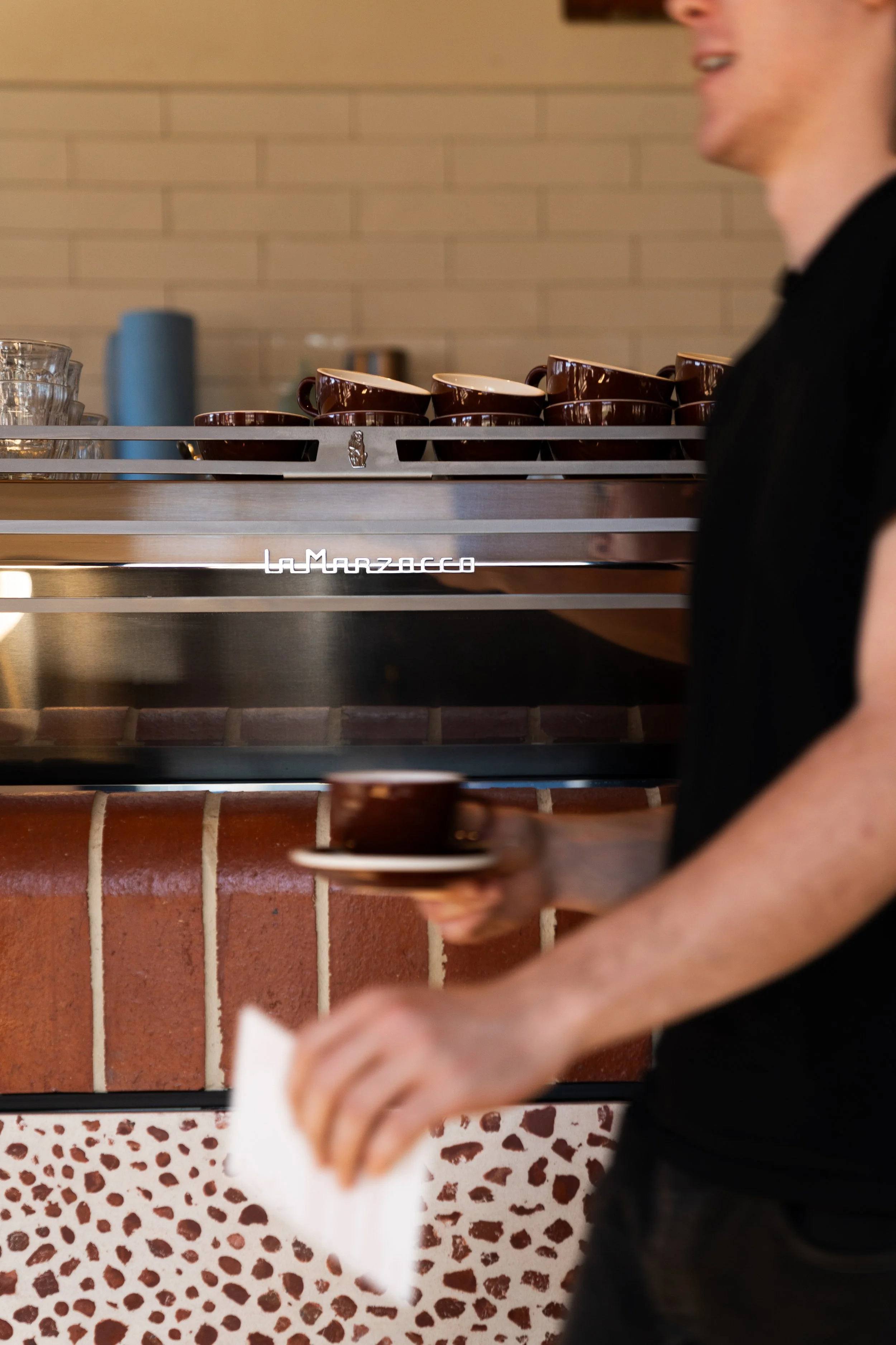 A person in black clothing holding a white napkin, standing in front of a counter with stacked brown coffee cups on a silver tray, a row of glasses, and a brick wall background.