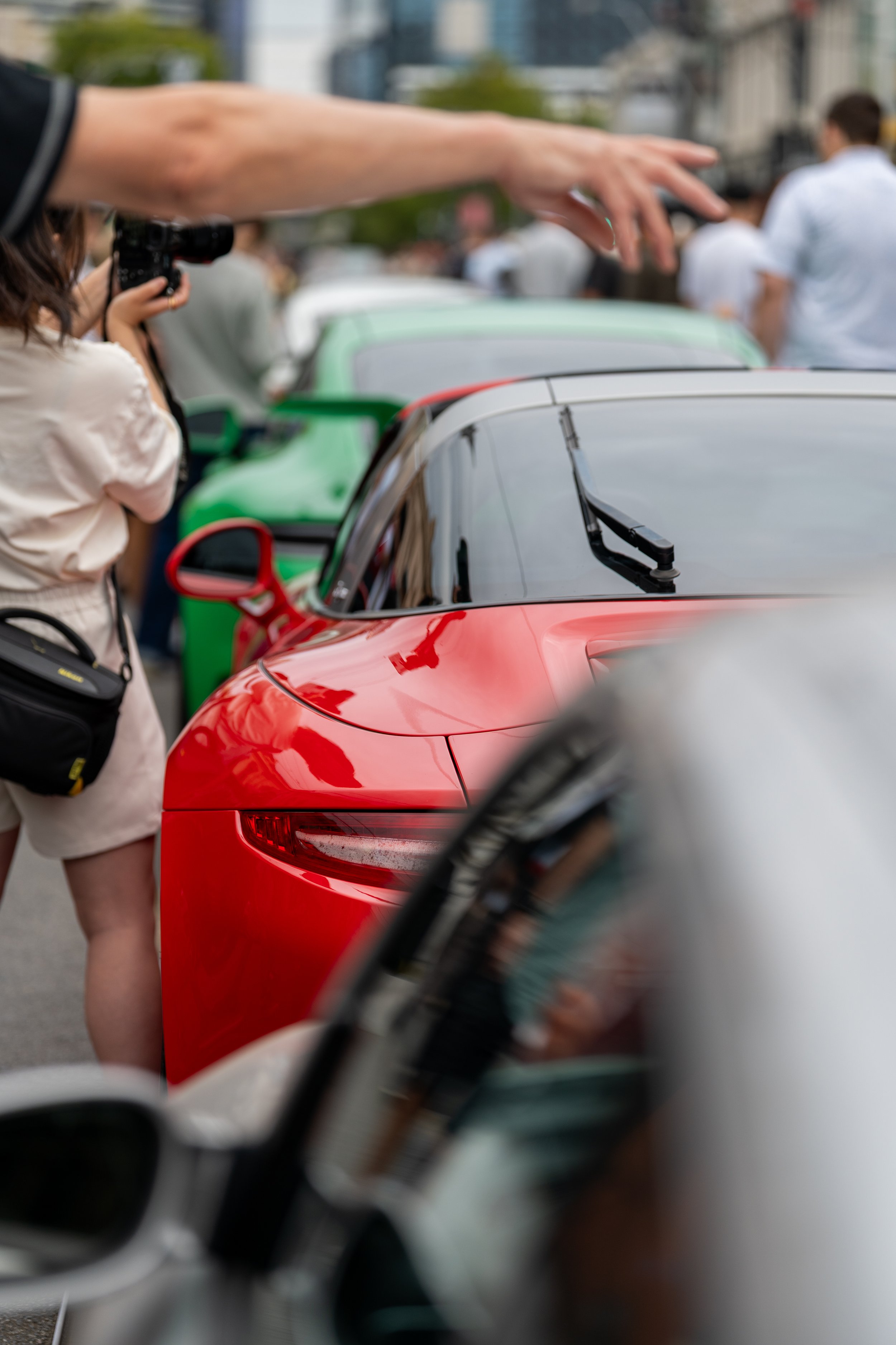 A person in a white shirt taking a picture with a camera on a busy street with cars and people in the background. Porsche 911 Car Meet in Melbourne australia motorsport photography
