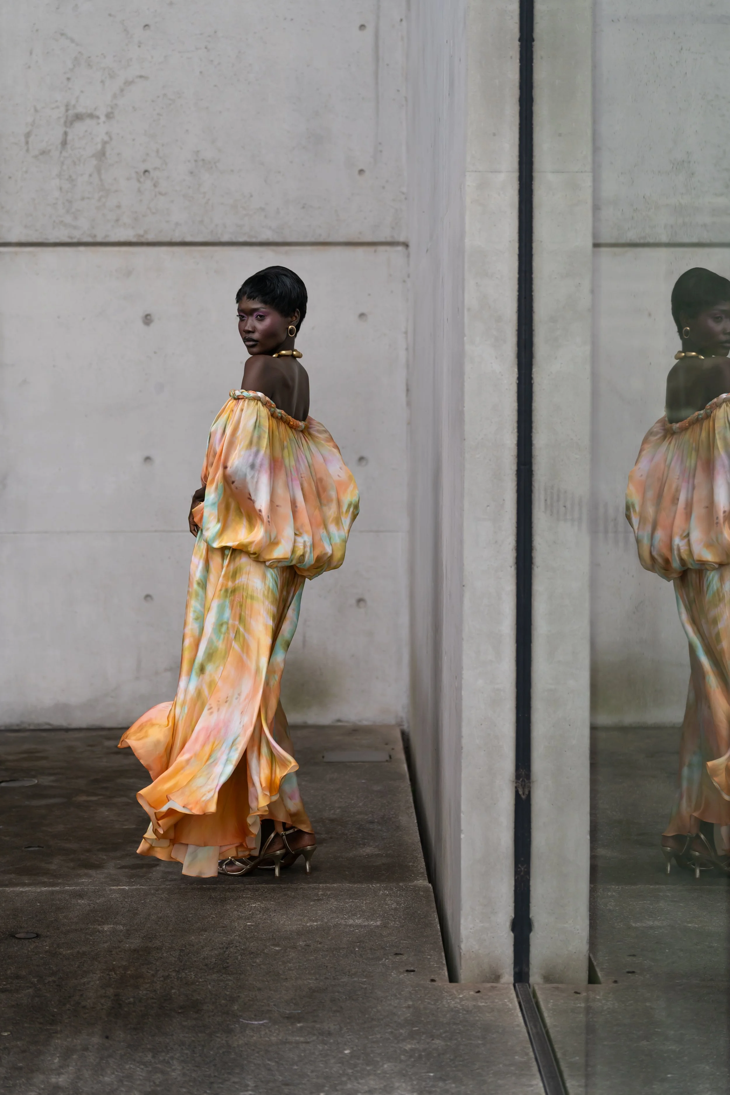A woman with short black hair and dark skin, wearing a colorful, flowing, off-the-shoulder dress with a pastel tie-dye pattern, stands against a concrete wall. She is accessorized with gold jewelry, including earrings, a necklace, and bracelets, and 