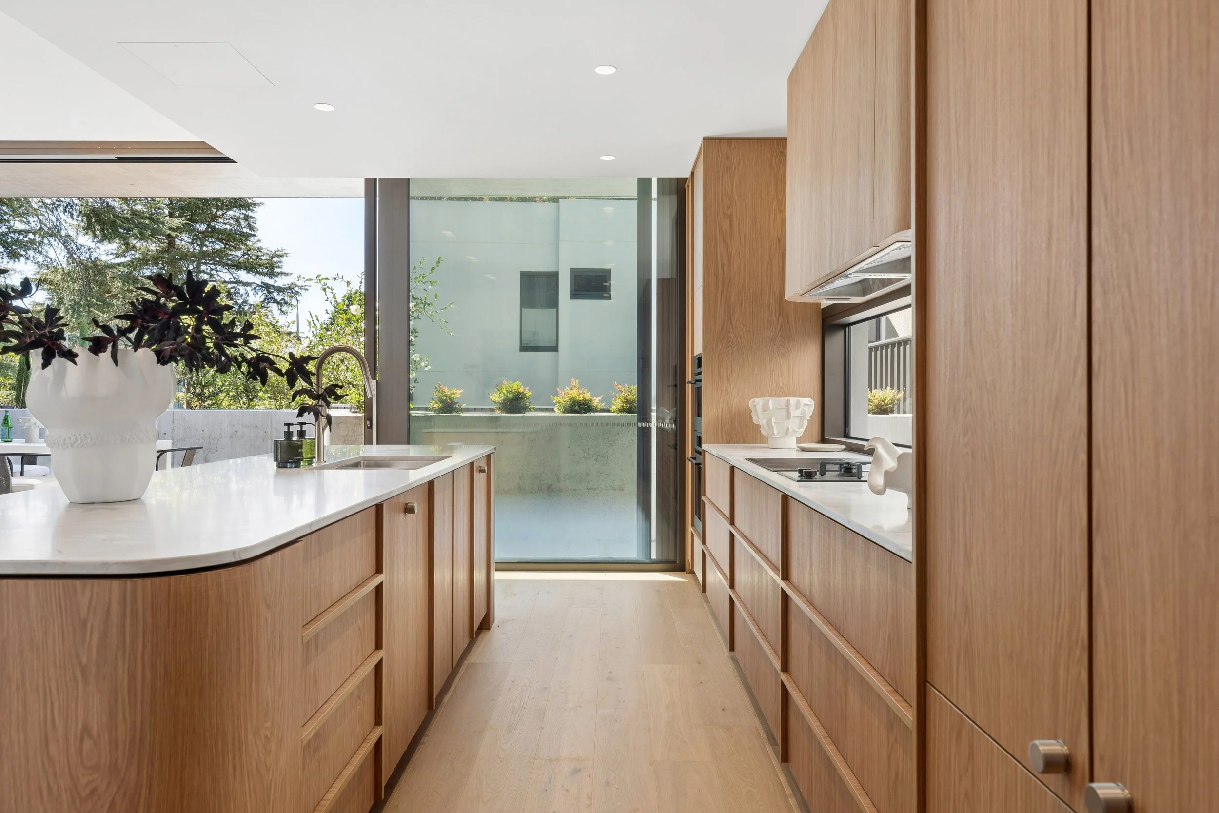 A modern kitchen with wooden cabinetry, white countertops, a vase with dark purple flowers, and large windows bringing in natural light. architecturally designed house in forrest canberra, editorial photography