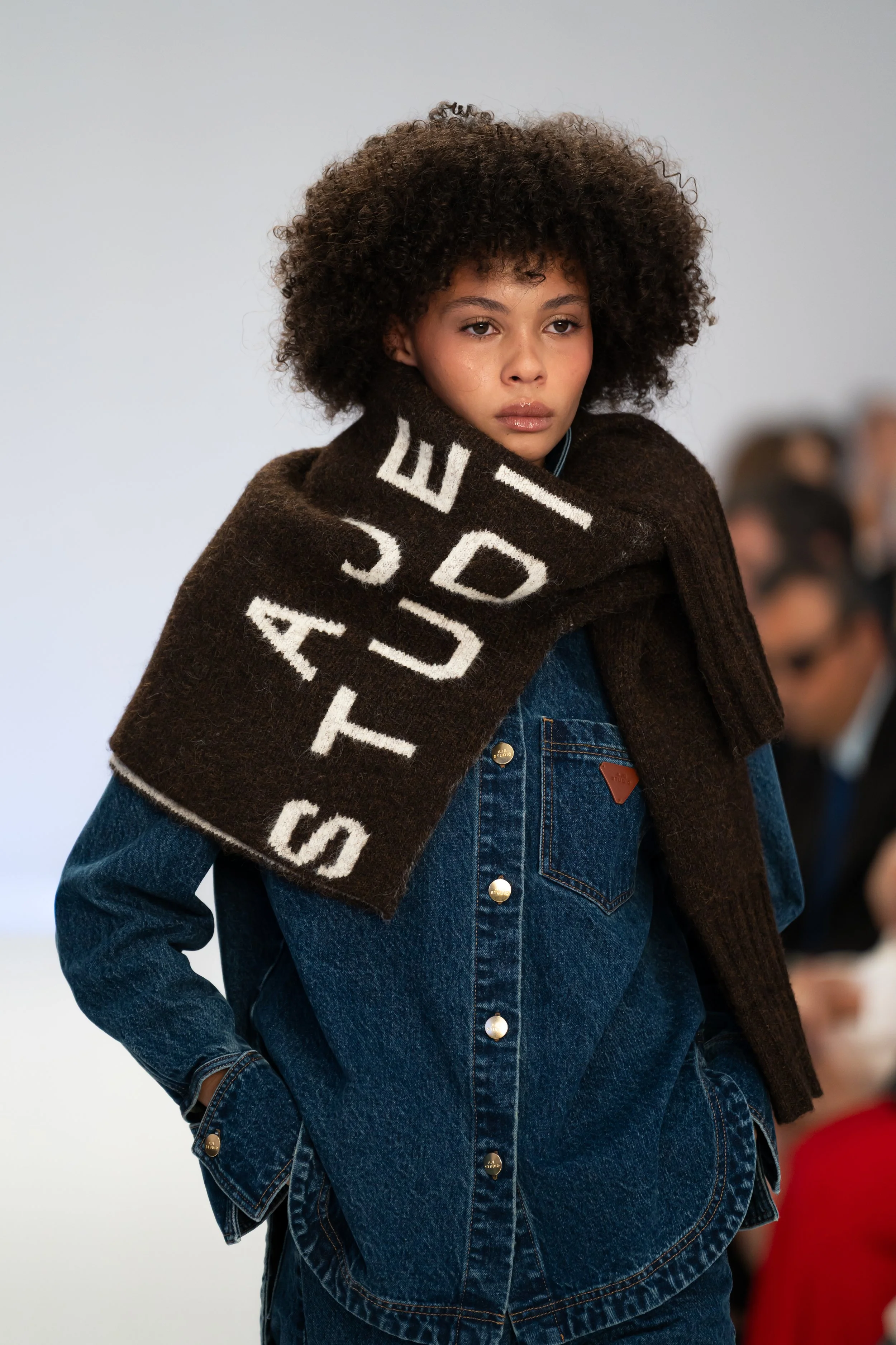 A young woman with curly hair wearing a brown shawl with white letters and a denim jacket, standing indoors at a fashion show runway. Australian fashion week for Aje studio