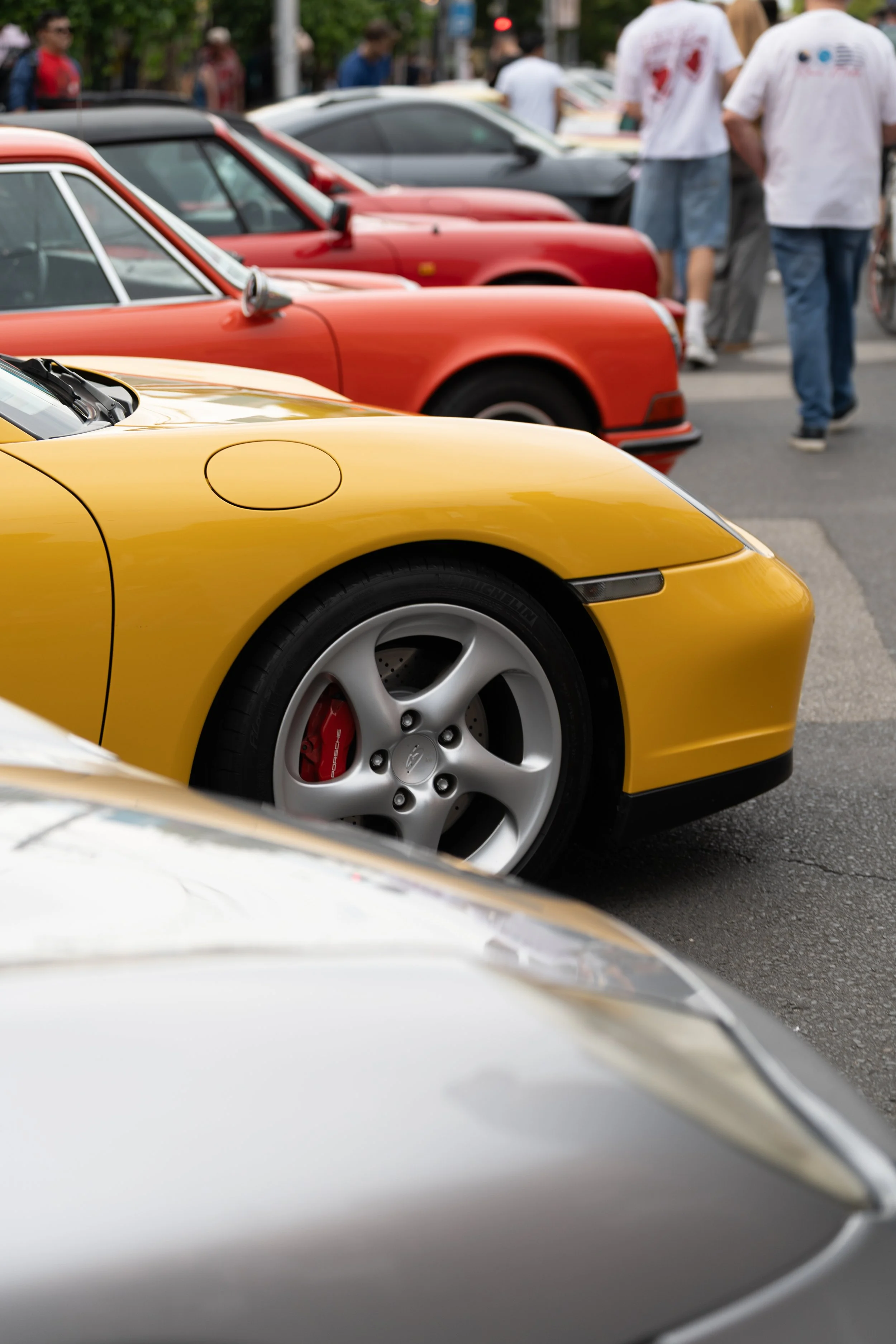 Close-up of a yellow sports car Porsche 911 Carrera parked at a car show, with other colorful cars and people in the background. motorsport street photography