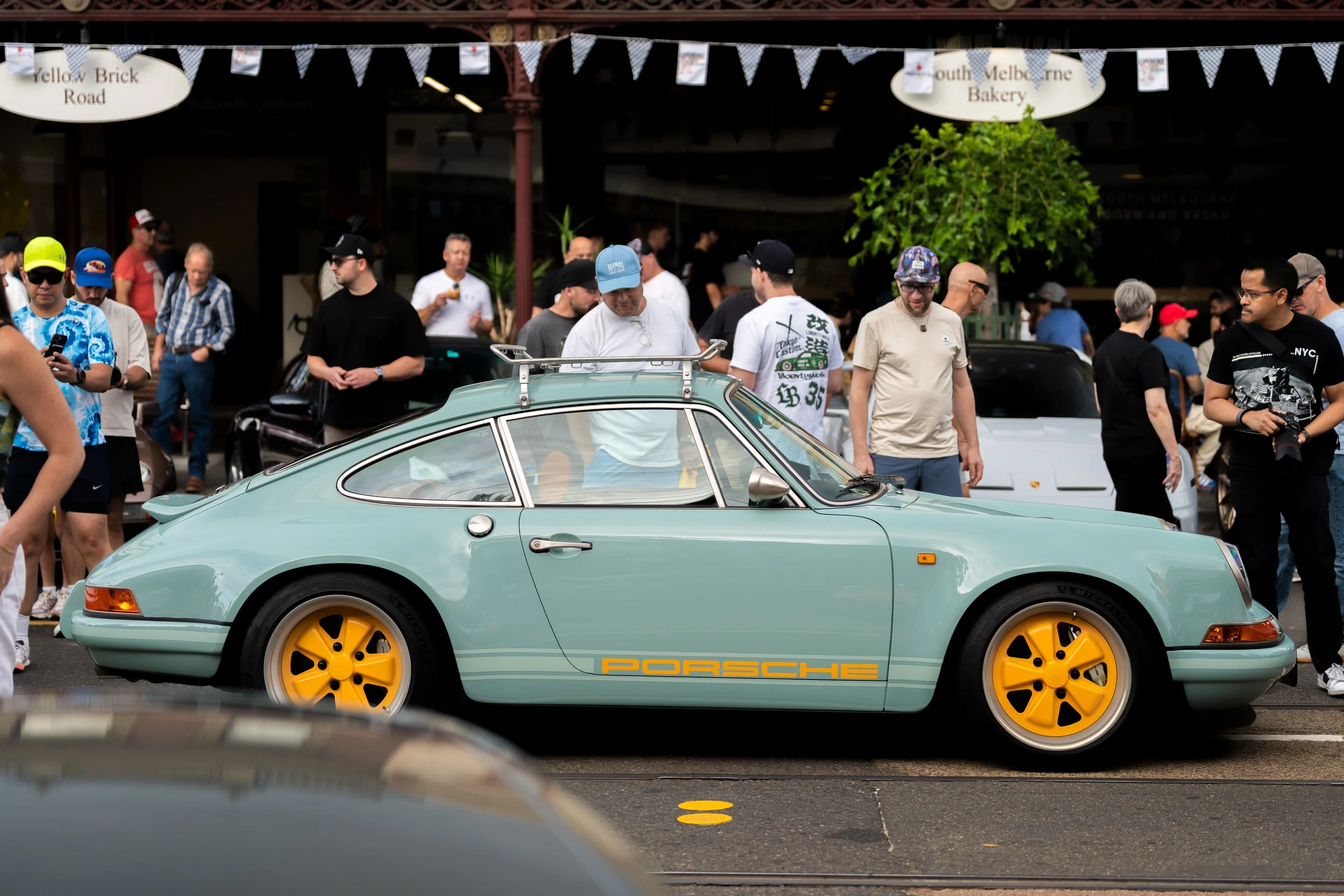 A crowd of people gathered around a light blue classic Porsche car with yellow wheels, at an outdoor car event. The background features signage for a bakery and a street name, with banners and trees. Vintage Porsche 911
