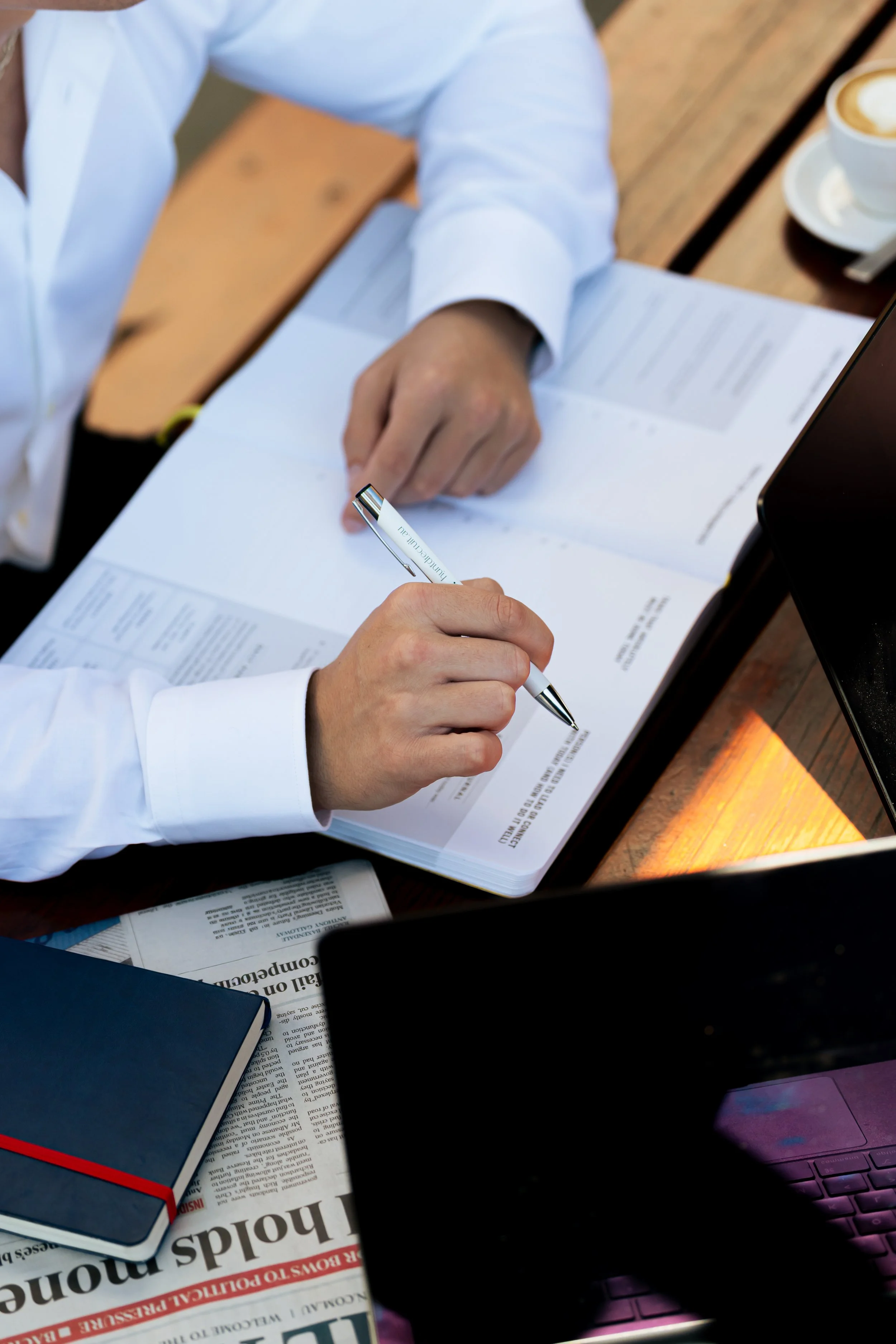 A person in a white shirt is sitting at a wooden table, writing on a document with a silver pen. On the table, there are newspapers, a blue notebook with a red elastic band, and a cup of coffee.