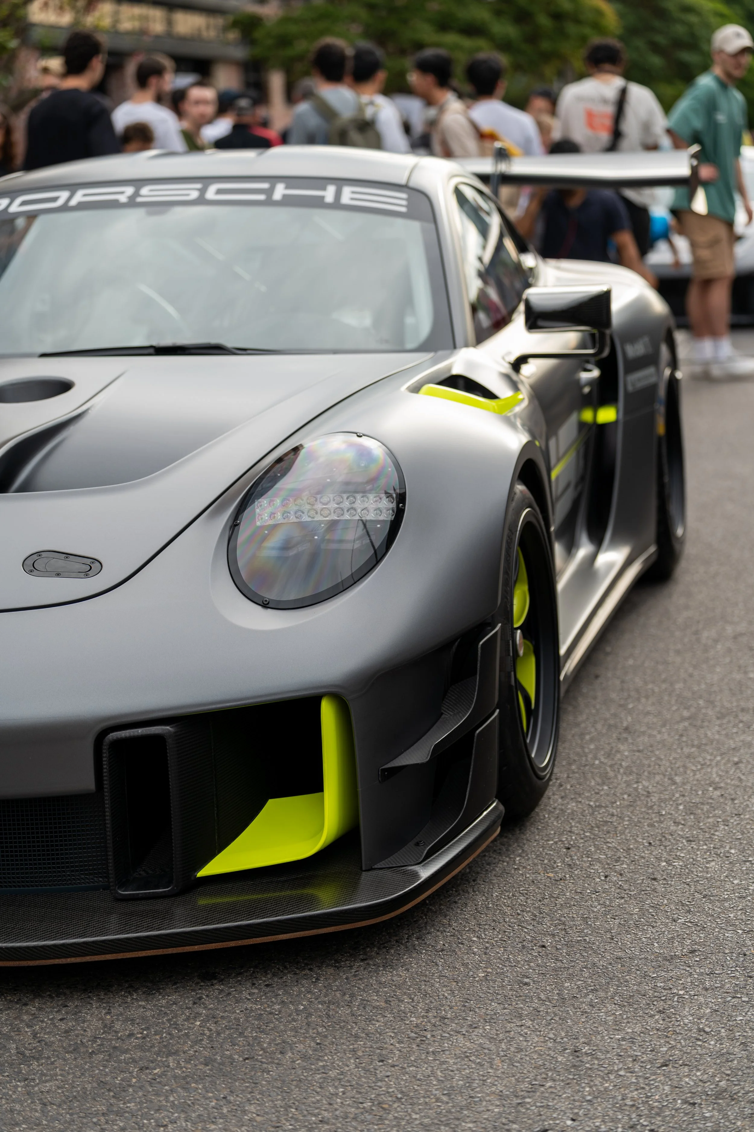 Close-up of a silver race car with green accents and Porsche branding, parked on a street with a crowd of people in the background. Porsche 911 gt3 rs carrera cup race car matte grey custom wrap after market 