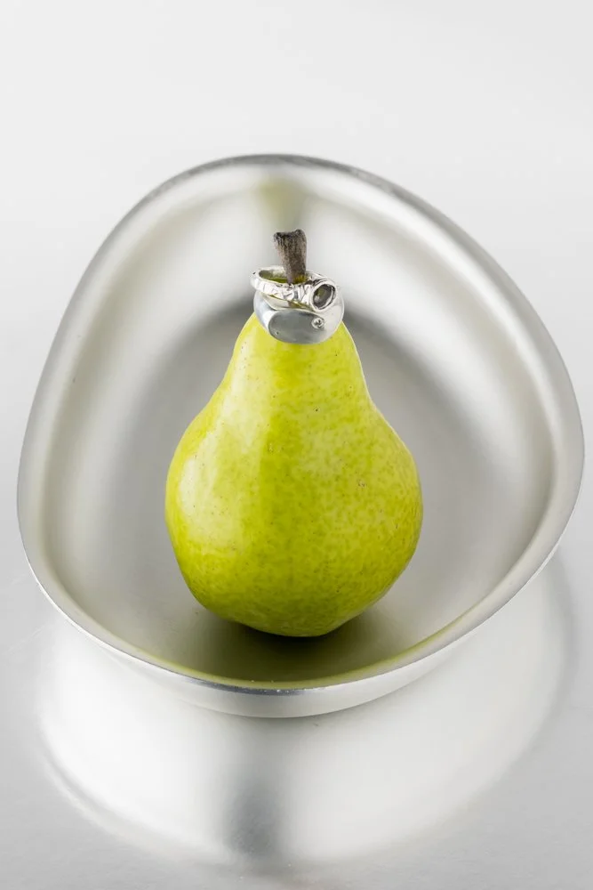 A green pear with a ring shaped like a snake around its top, placed in a white dish. fink dish  jewellery product photography