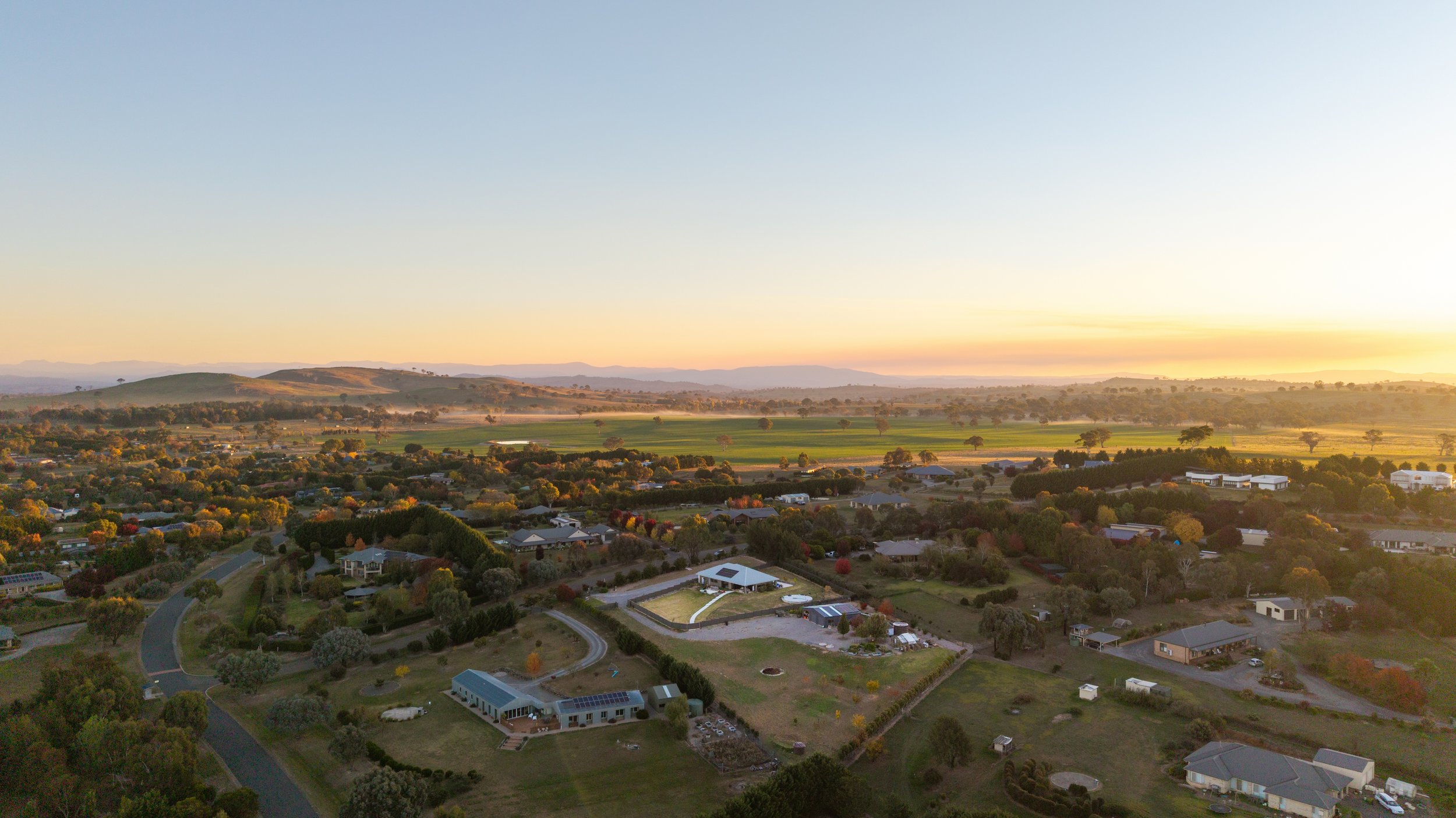 An aerial view of a suburban neighborhood with scattered houses, green lawns, and trees, with rolling hills in the background, during sunset. drone photography in murrumbateman real estate listing