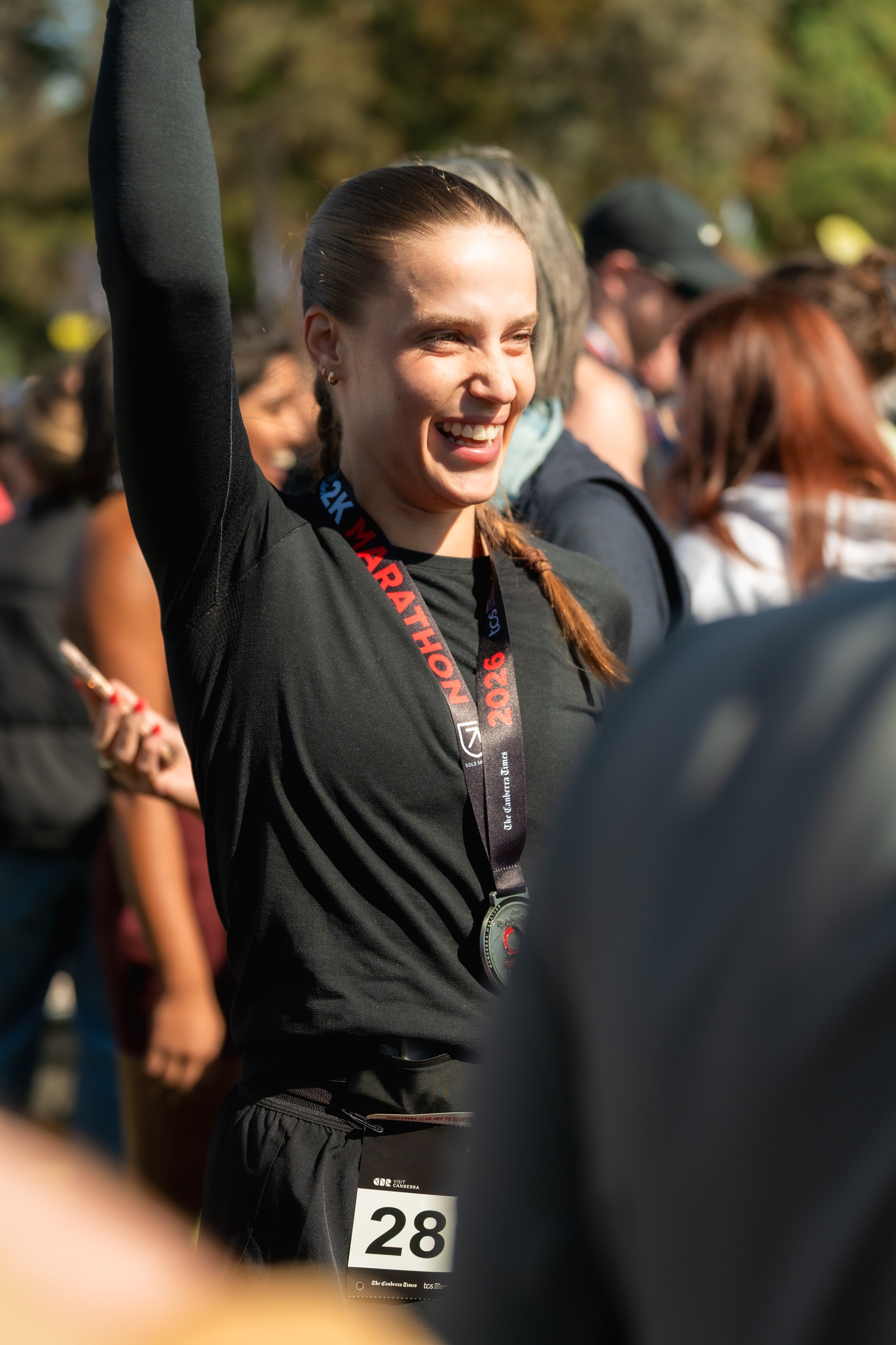A smiling woman with braided hair wearing a black athletic shirt and a medal around her neck at an outdoor event, with other people in the background. Canberra marathon finisher 2026 sports photography