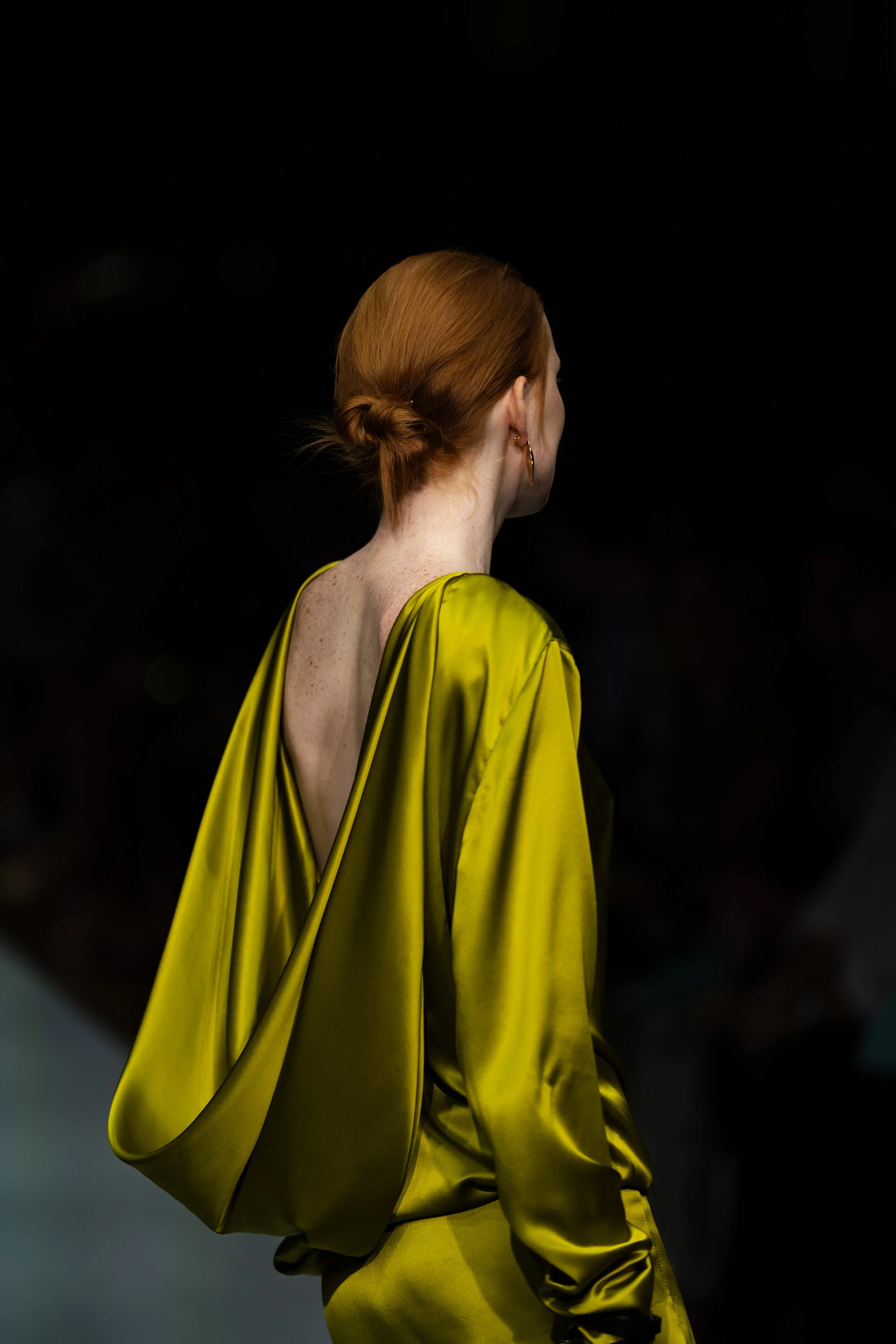 Side view of a woman with red hair styled in a low bun, wearing gold hoop earrings and a loose-fitting yellow satin dress with an open back, against a dark background. melbourne fashion festival cappallazzo runway show