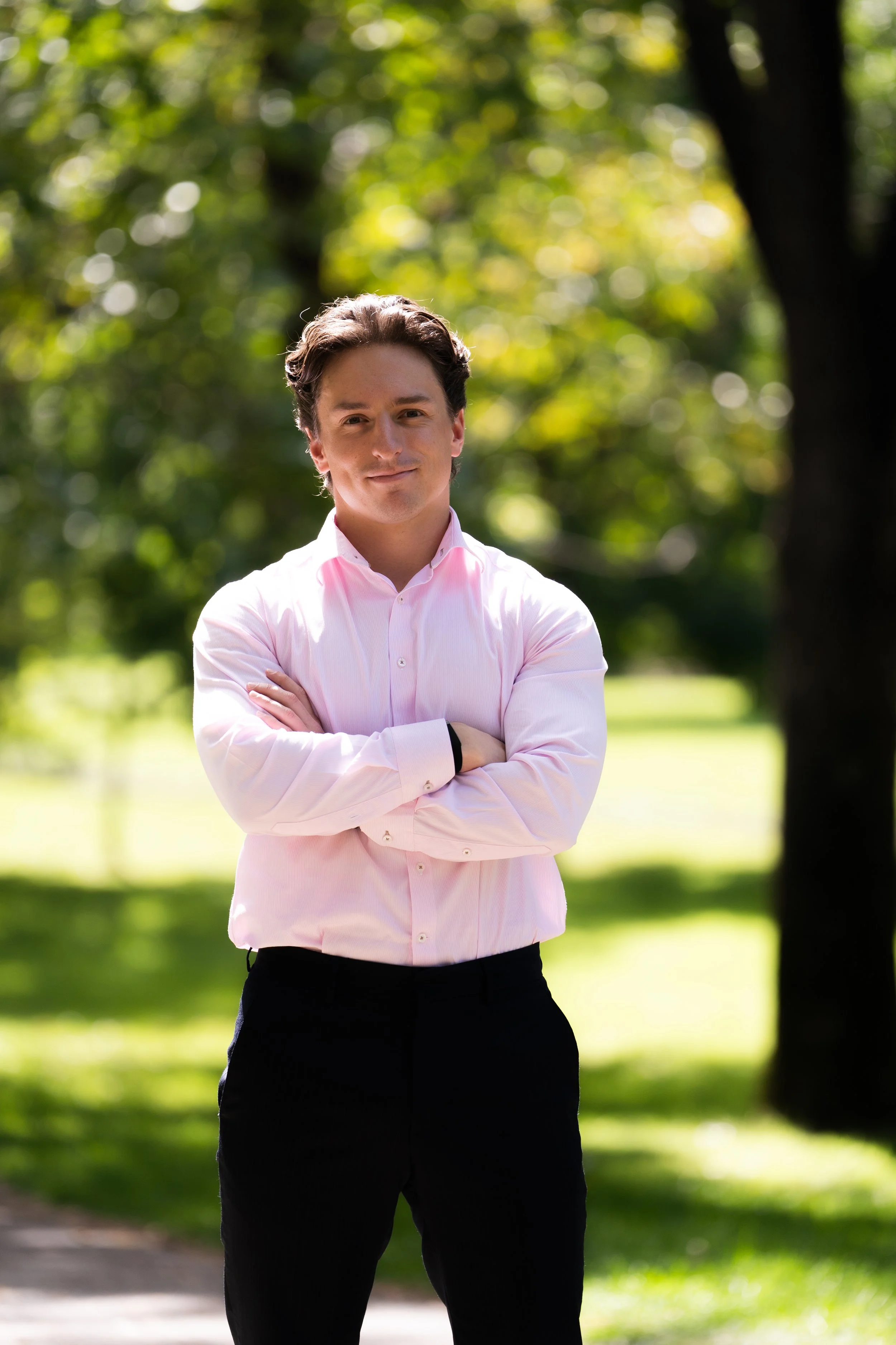 A young man with dark hair, wearing a pink collared shirt, standing outdoors with arms crossed, in a park with green trees and sunlight. Financial professional headshot 