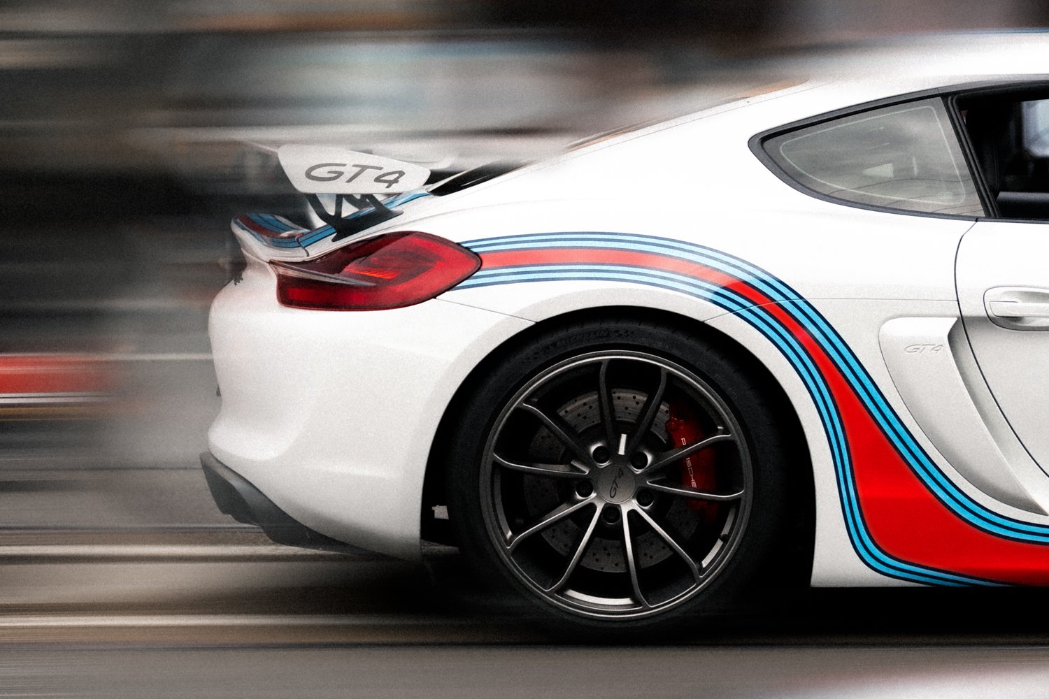 A white Porsche 911 GT4 race car with blue and red racing stripes, featuring a rear wing, black rims, a visible brake caliper, and a blurred background indicating motion.