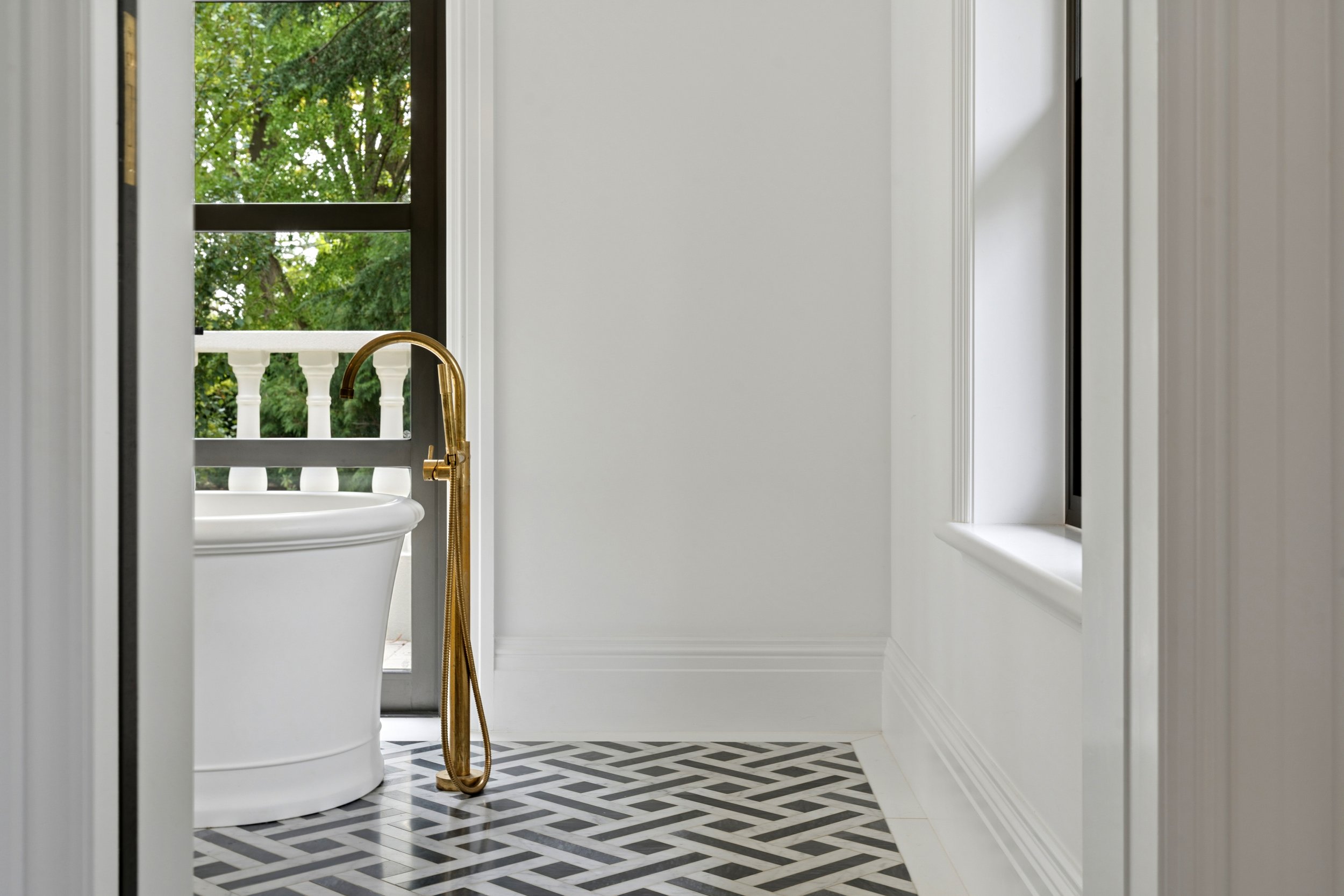 Bathroom with black and white geometric patterned tile floor, white walls, a window with trees outside, and a clawfoot bathtub with a gold faucet. editorial architectural photography Red Hill canberra interior designer Thomas Hamel