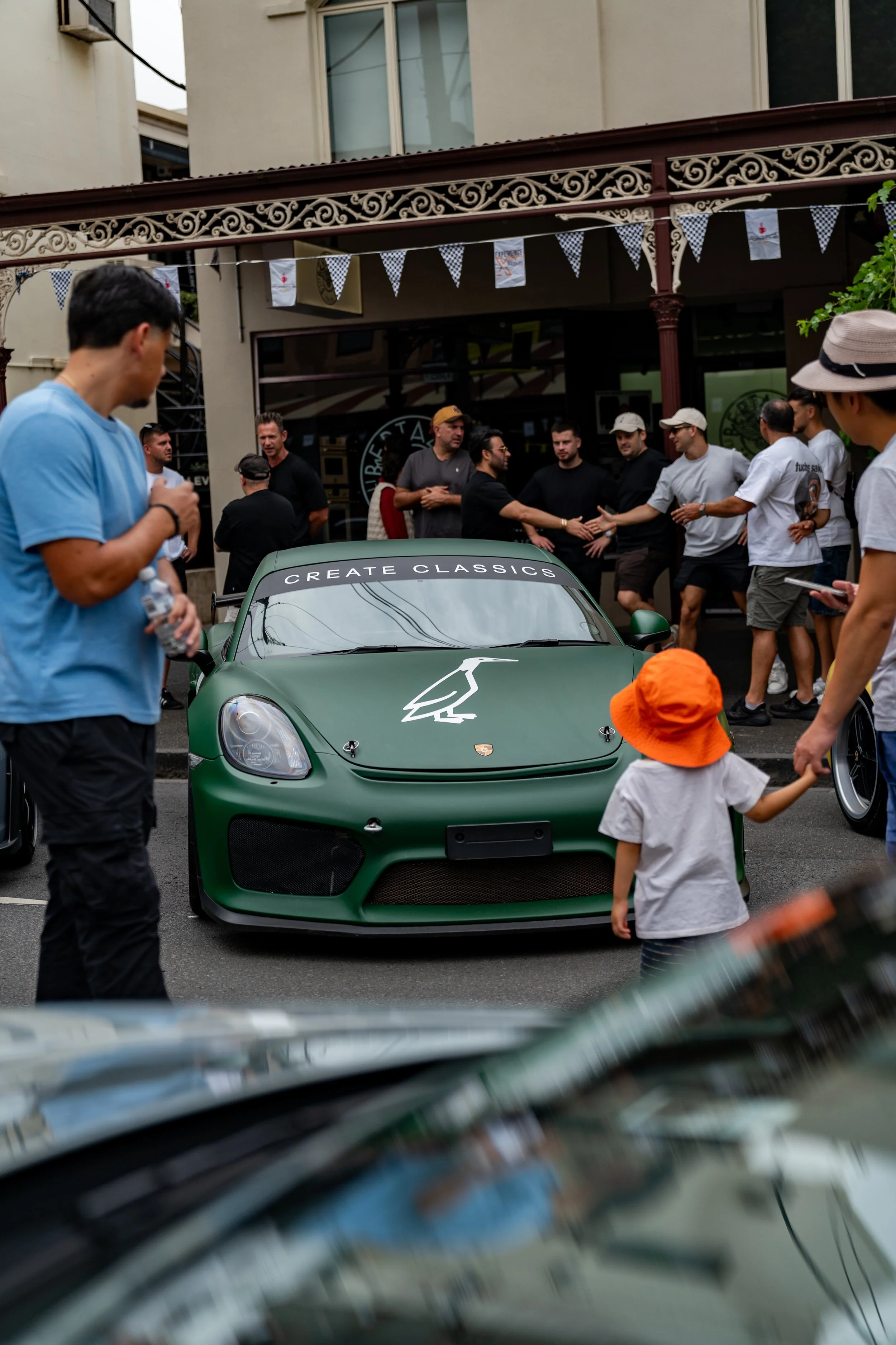 A green Porsche sports car with the phrase 'CREATE CLASSICS' on the windshield is parked in a city street. Several people are gathered around, some shaking hands, others talking or walking. The scene appears to be part of a car event or gathering.