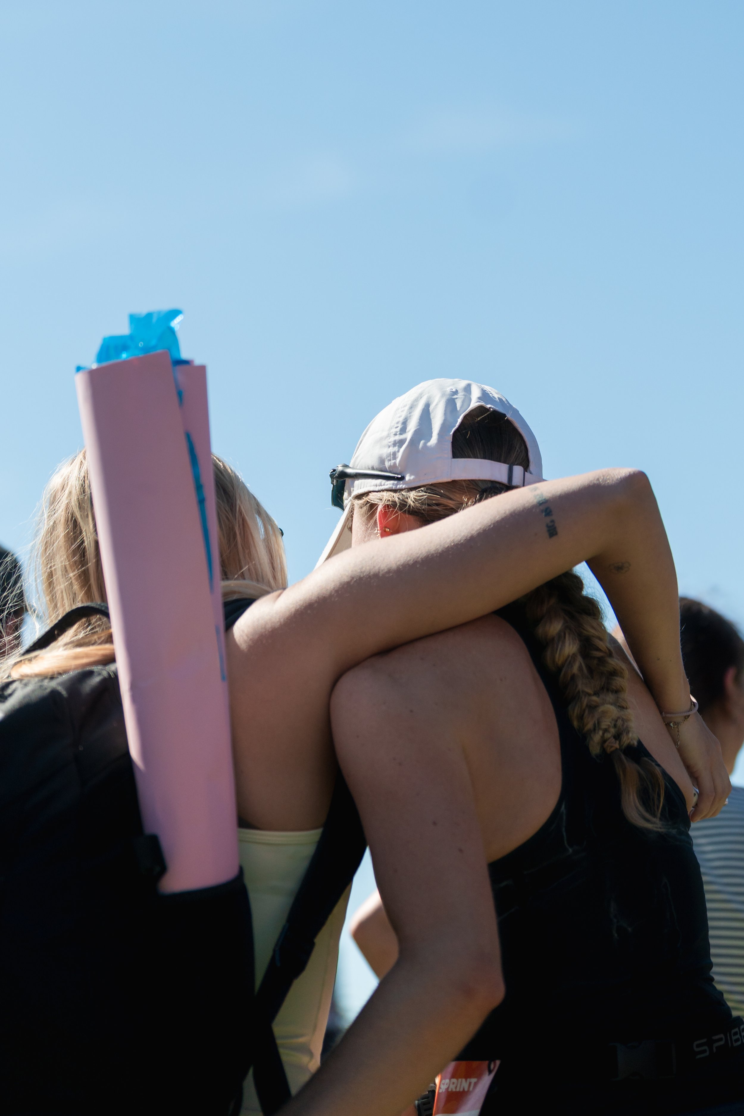 A group of people with their arms around each other's shoulders, wearing athletic clothing and backpacks, outdoors on a sunny day with a clear blue sky. female friendship at Wollongong Triathlon 2026 sports photography