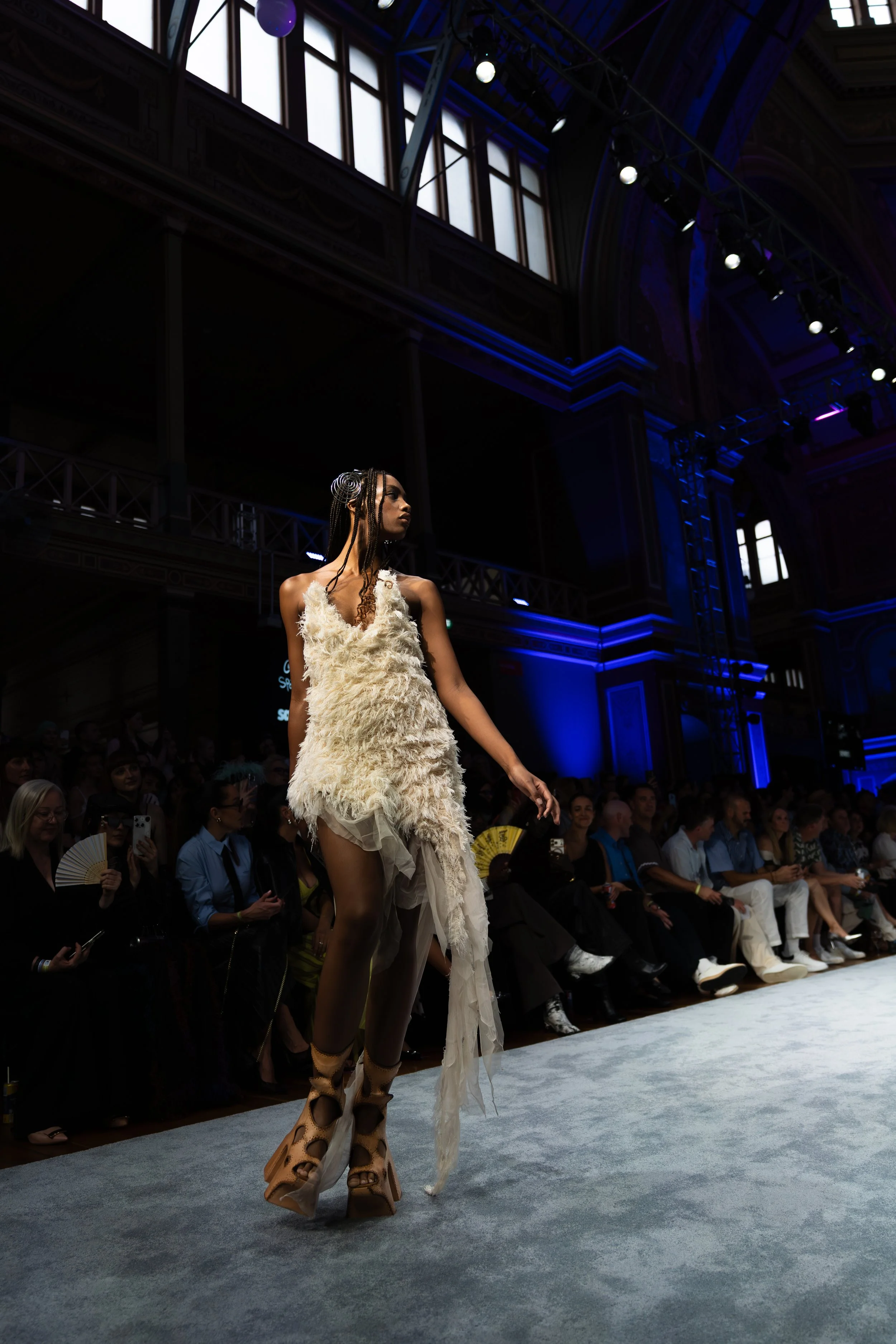 A model walking down a runway at a fashion show, wearing a cream-colored fuzzy dress over stockings and platform shoes. The runway is in a grand hall with stained glass windows and an audience seated on either side. melbourne fashion festival