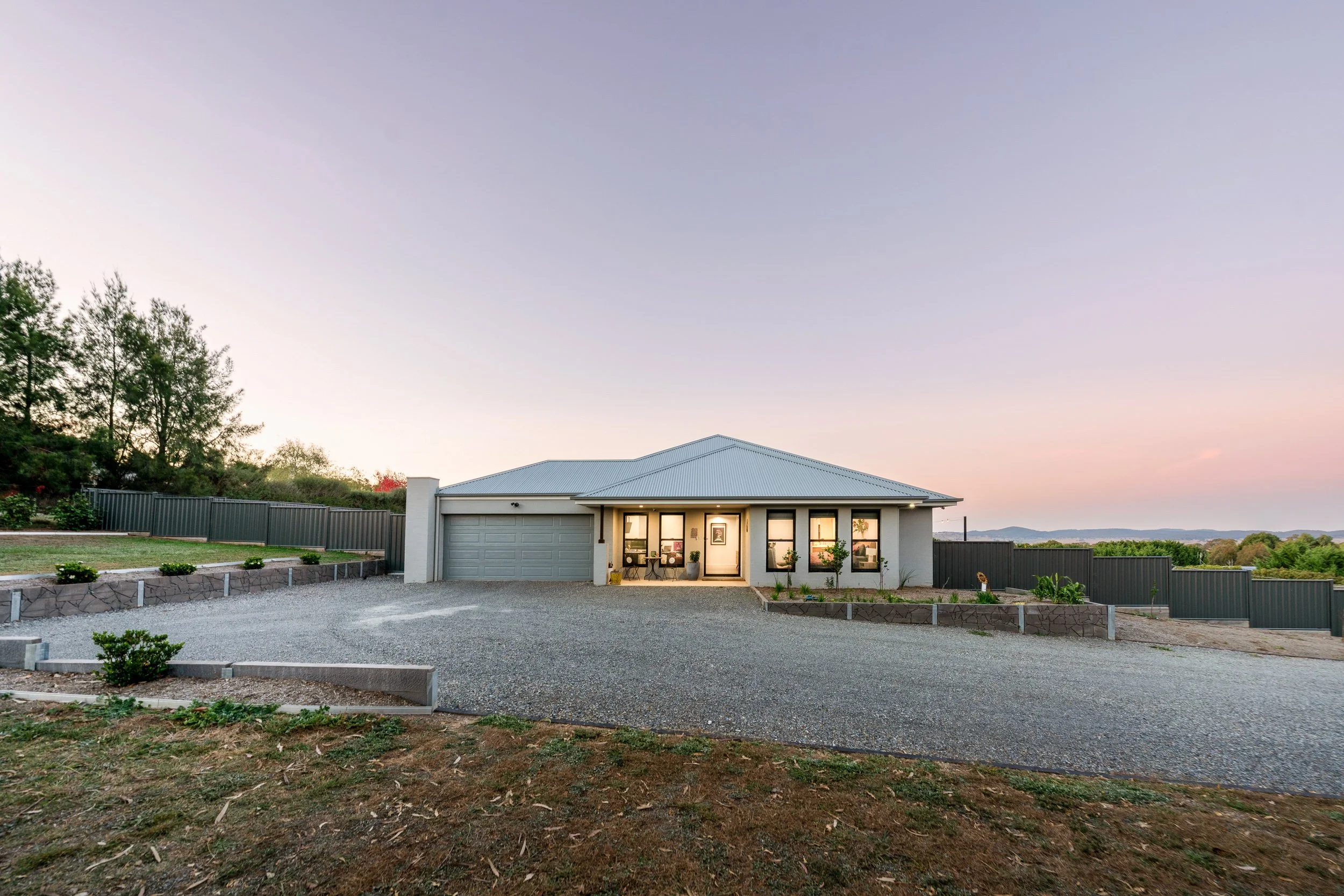 Front view of a modern house at dusk with a well-lit porch and large windows, a gravel driveway, and a landscaped yard. Murrumbateman real estate dusk twilight photography
