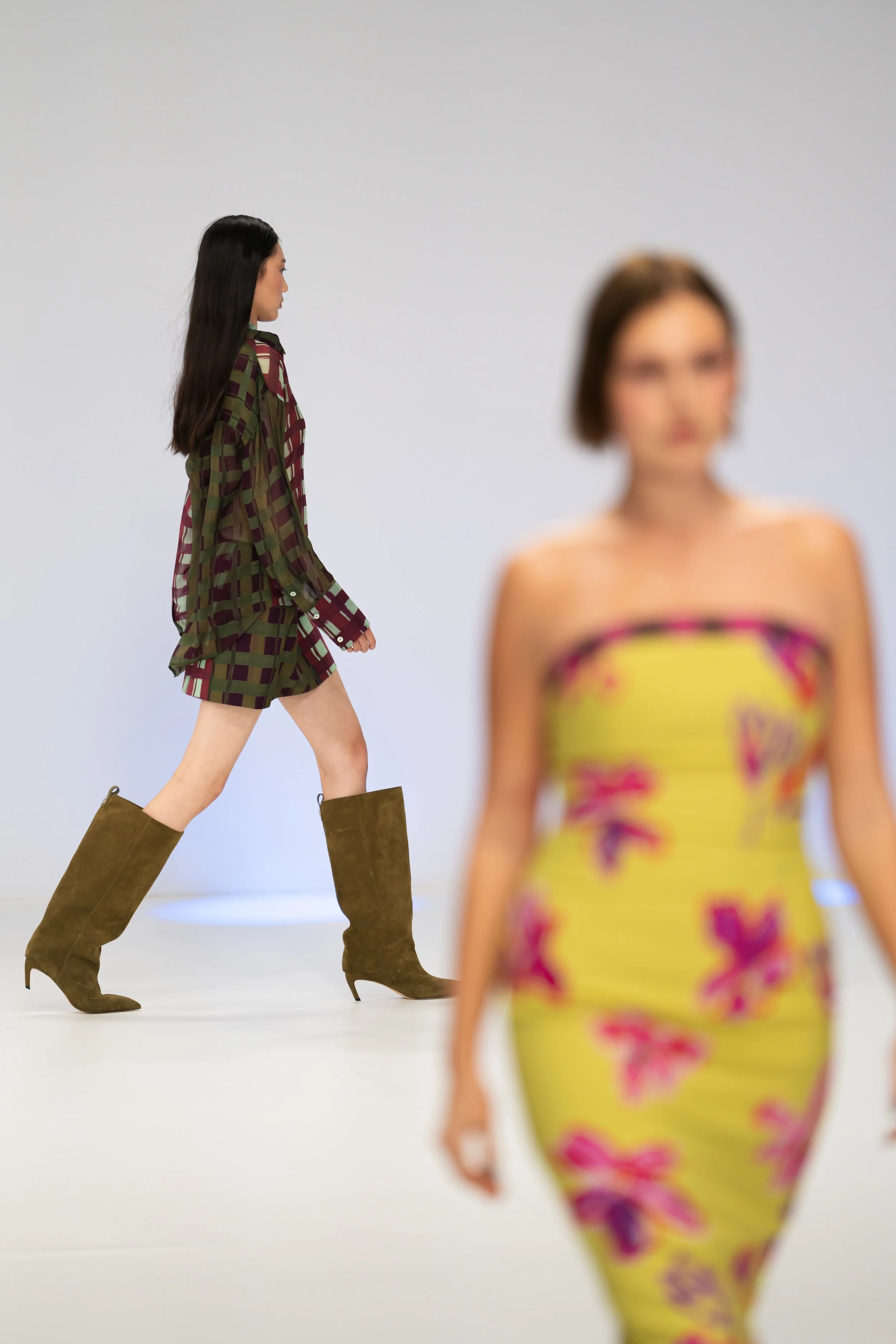 A woman with long black hair dressed in a checkered green and red dress walking in front of another woman with short hair in a yellow dress with purple and pink pattern, against a plain white background. australian fashion week runway photography